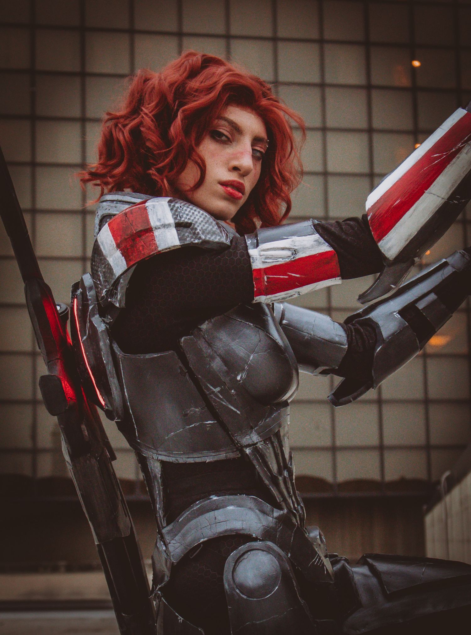 Red-haired cosplayer in detailed dark armor costume with red accents poses dramatically at a convention.