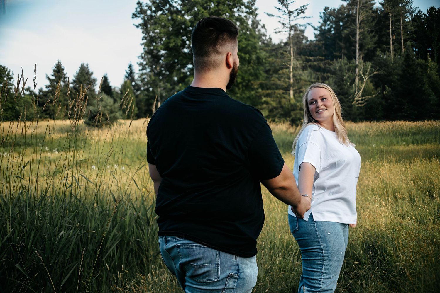 A couple shares an intimate moment in a sunlit meadow surrounded by evergreen trees.