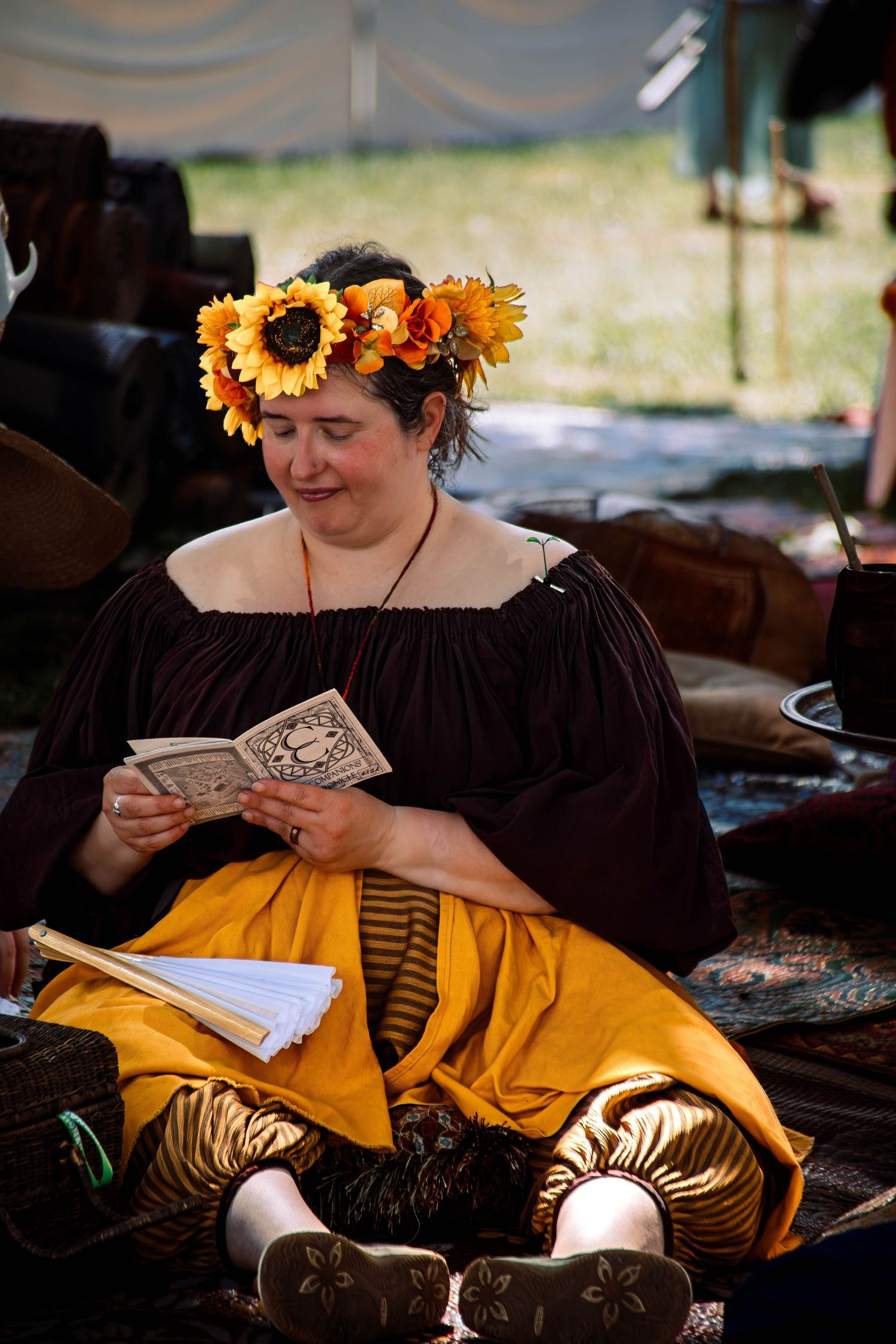Person in medieval attire wearing a sunflower crown reads from a book while sitting on yellow cushions.