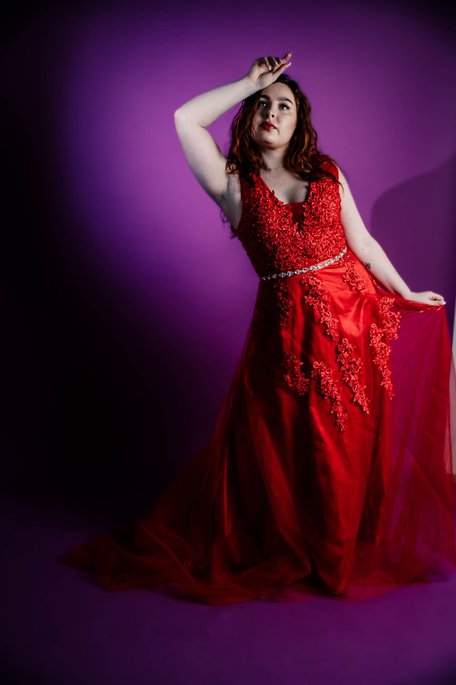 Woman in elegant red lace ballgown poses dramatically against purple background.