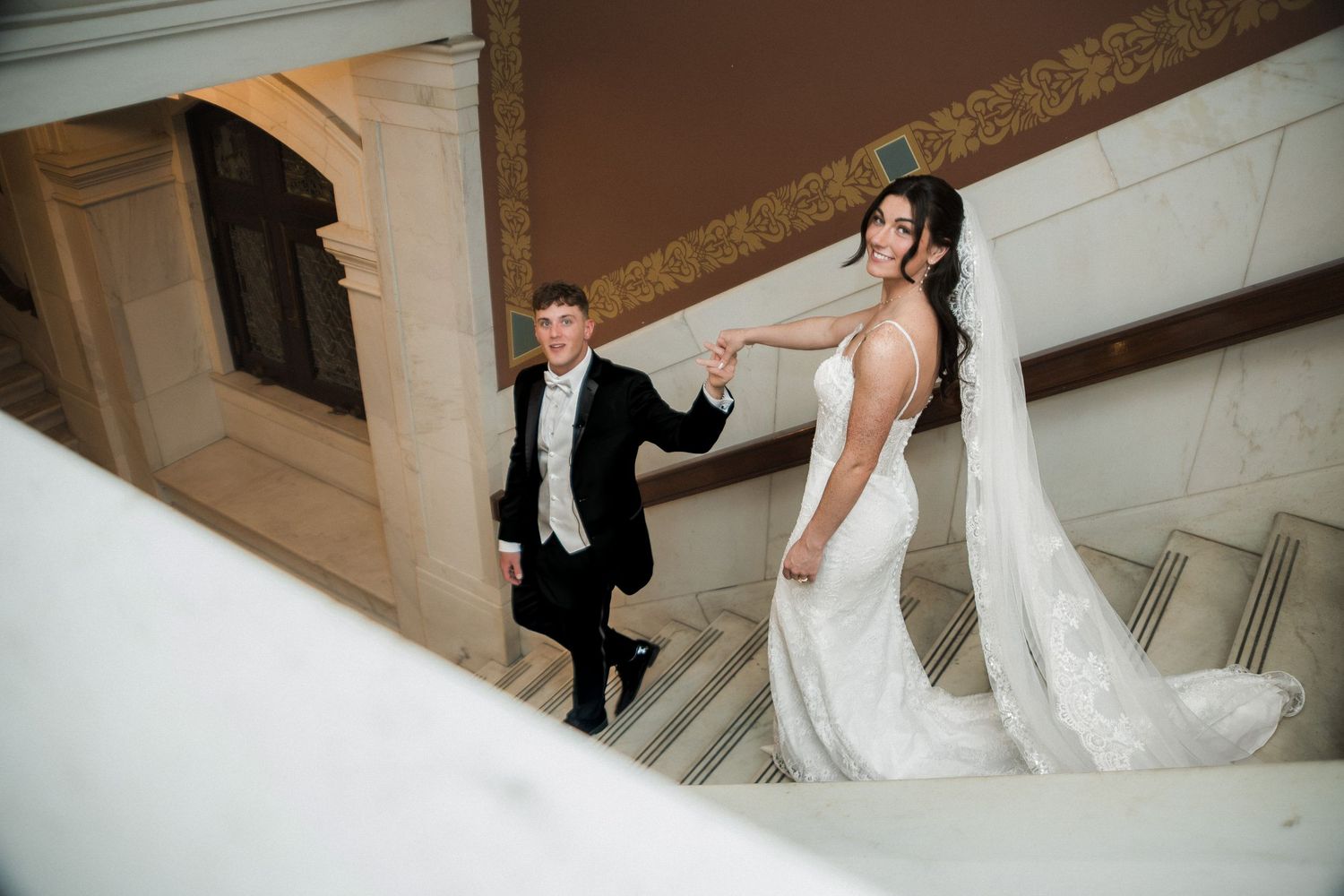 Bride and groom holding hands on elegant marble staircase, bride in lace mermaid gown with flowing train.