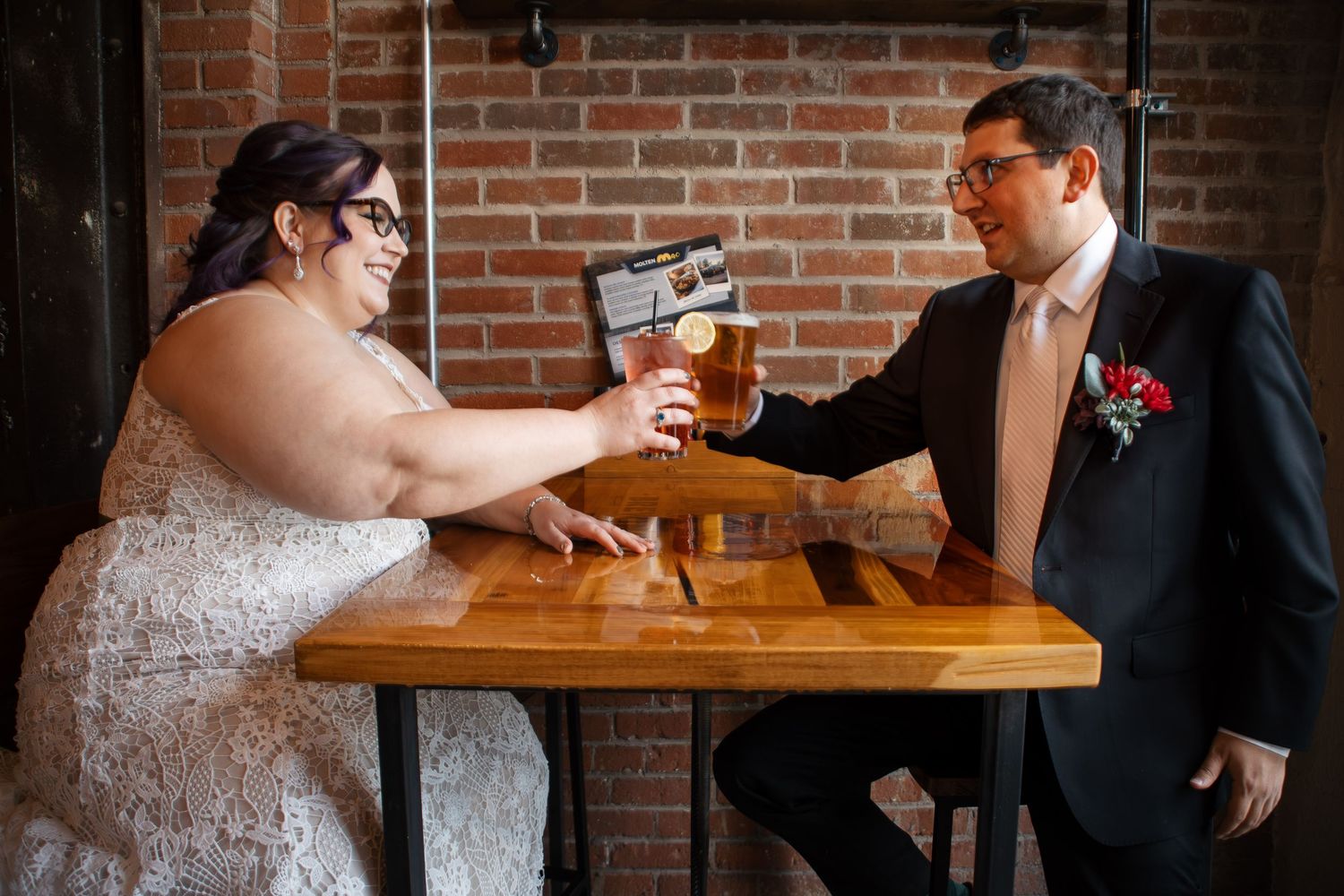 Bride and groom in wedding attire toasting with beer glasses at a wooden table in a brick-walled venue.