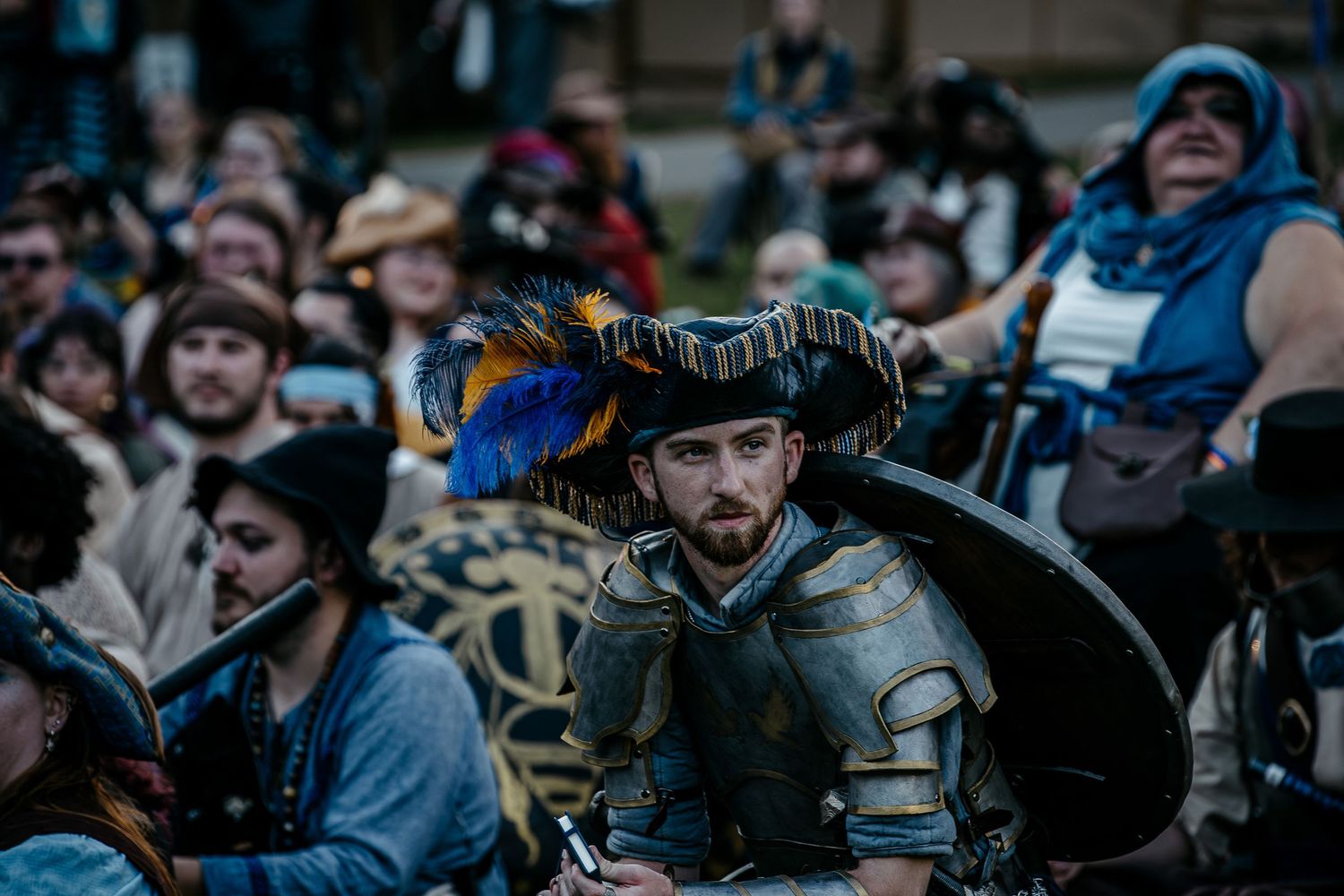 Man in Renaissance armor and feathered hat at a medieval fair, surrounded by costumed attendees in an outdoor setting.