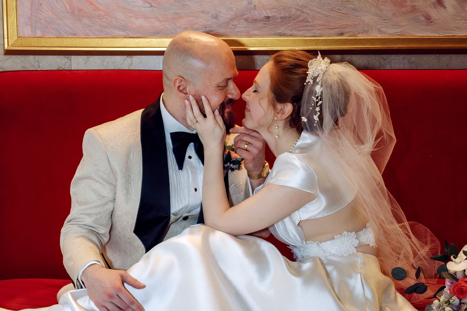 Bride and groom share a romantic kiss on a red velvet sofa during their wedding day celebration.