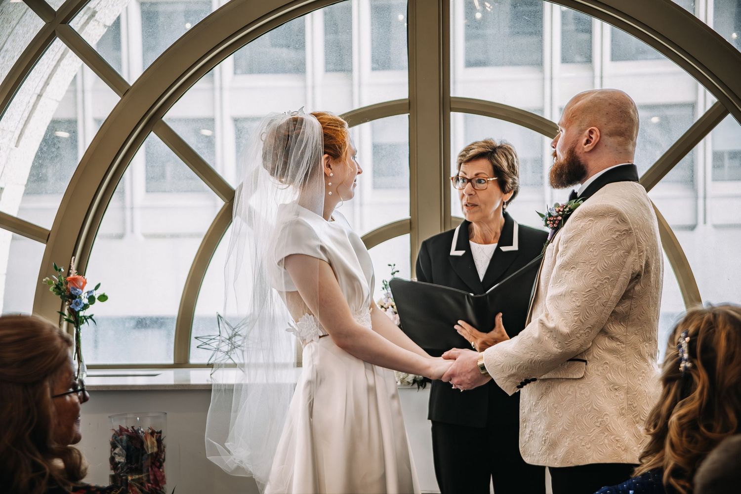 Bride and groom hold hands during indoor wedding ceremony with officiant before arched windows.
