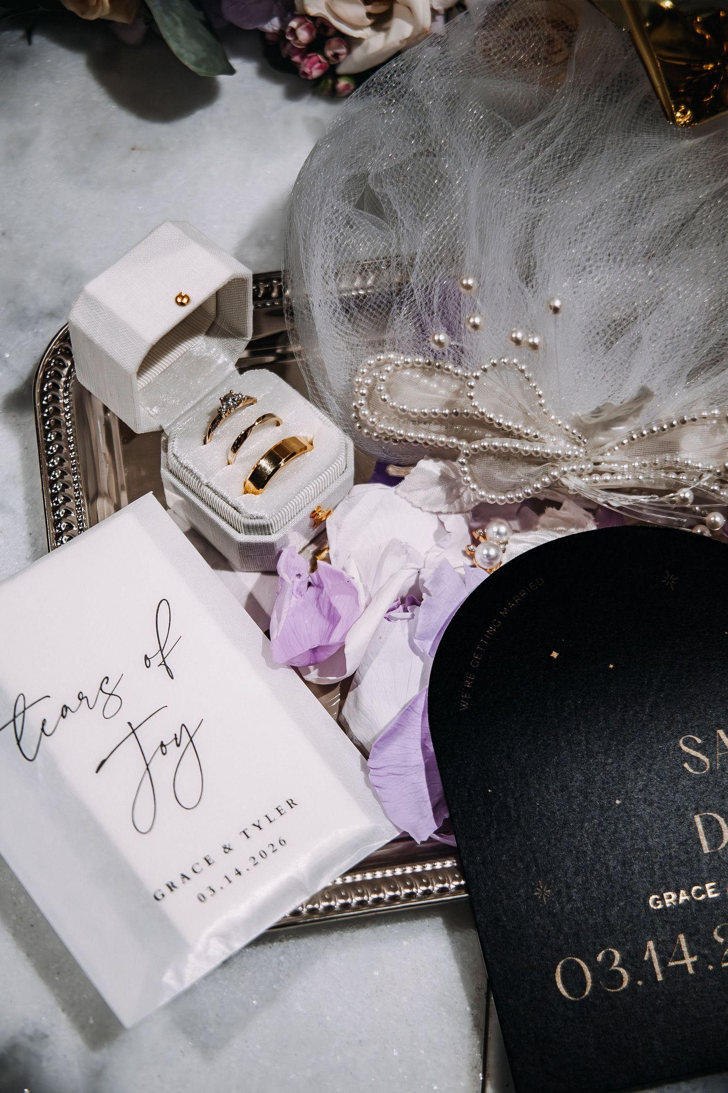 Wedding flat lay with gold rings, tears of joy packets, pearl necklace, veil, and black keepsake box dated 03.14.