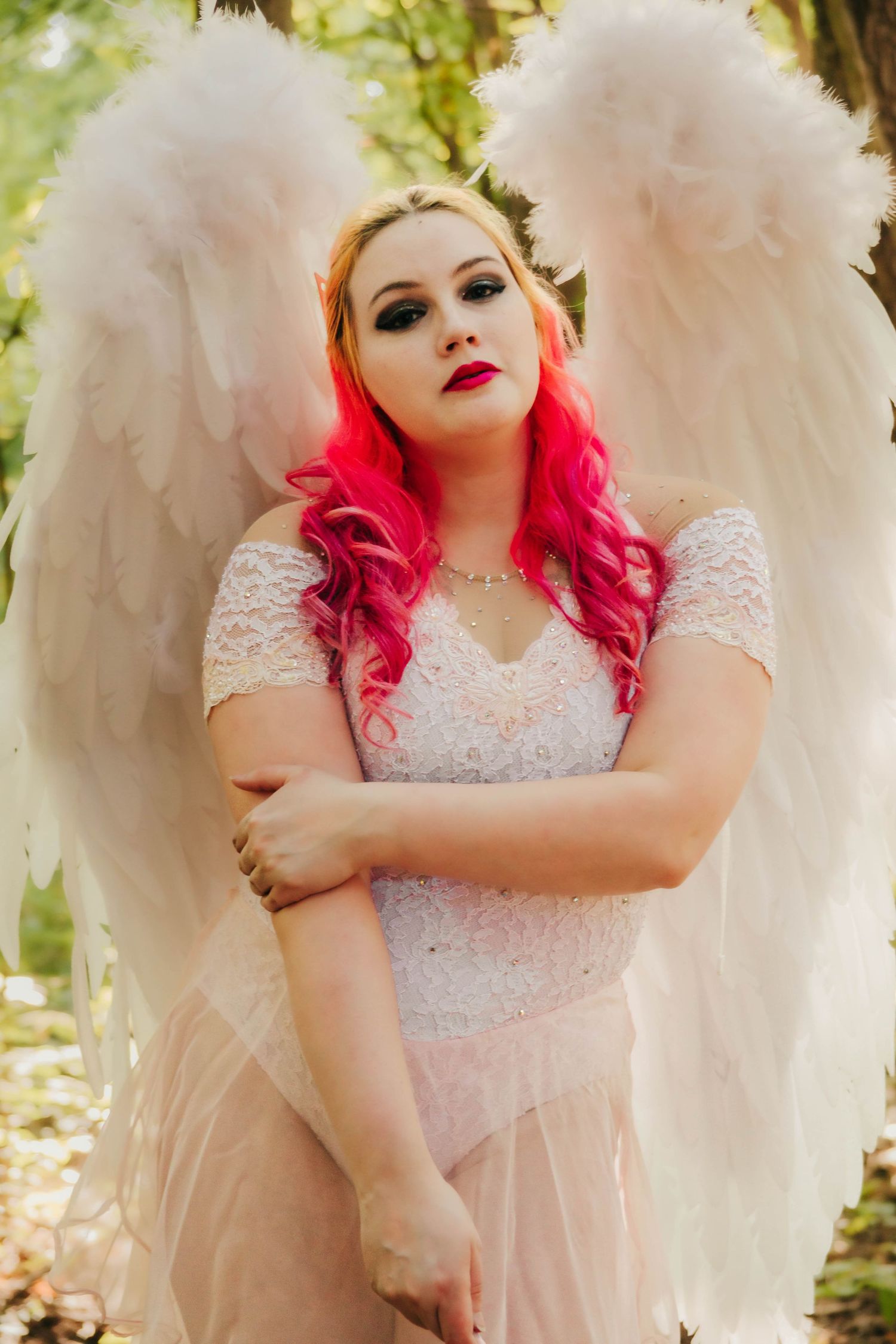 Woman with pink hair in angel costume with white feather wings wearing white lace dress in forest setting.