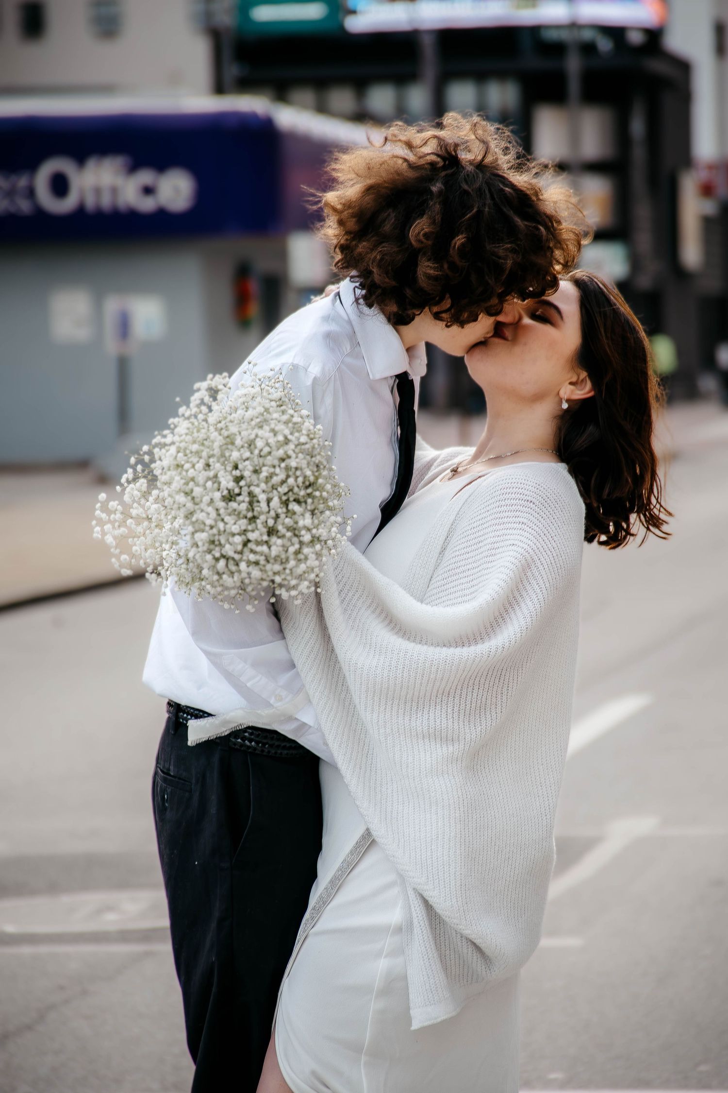 Couple kissing on a city street, woman in white sweater holding baby's breath bouquet, romantic urban elopement photo.