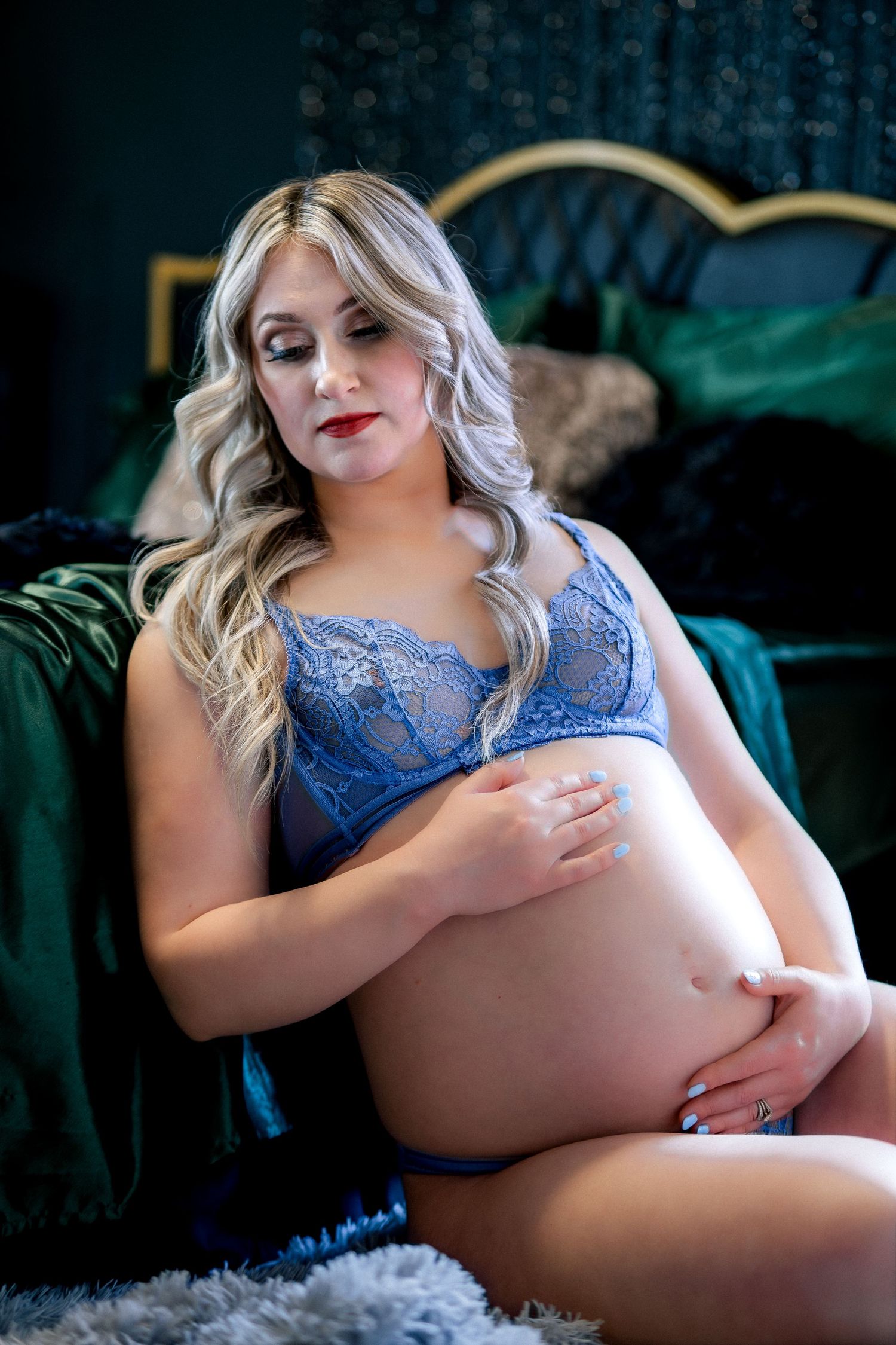 Pregnant woman in blue lace lingerie cradling her baby bump while sitting near a green velvet headboard.