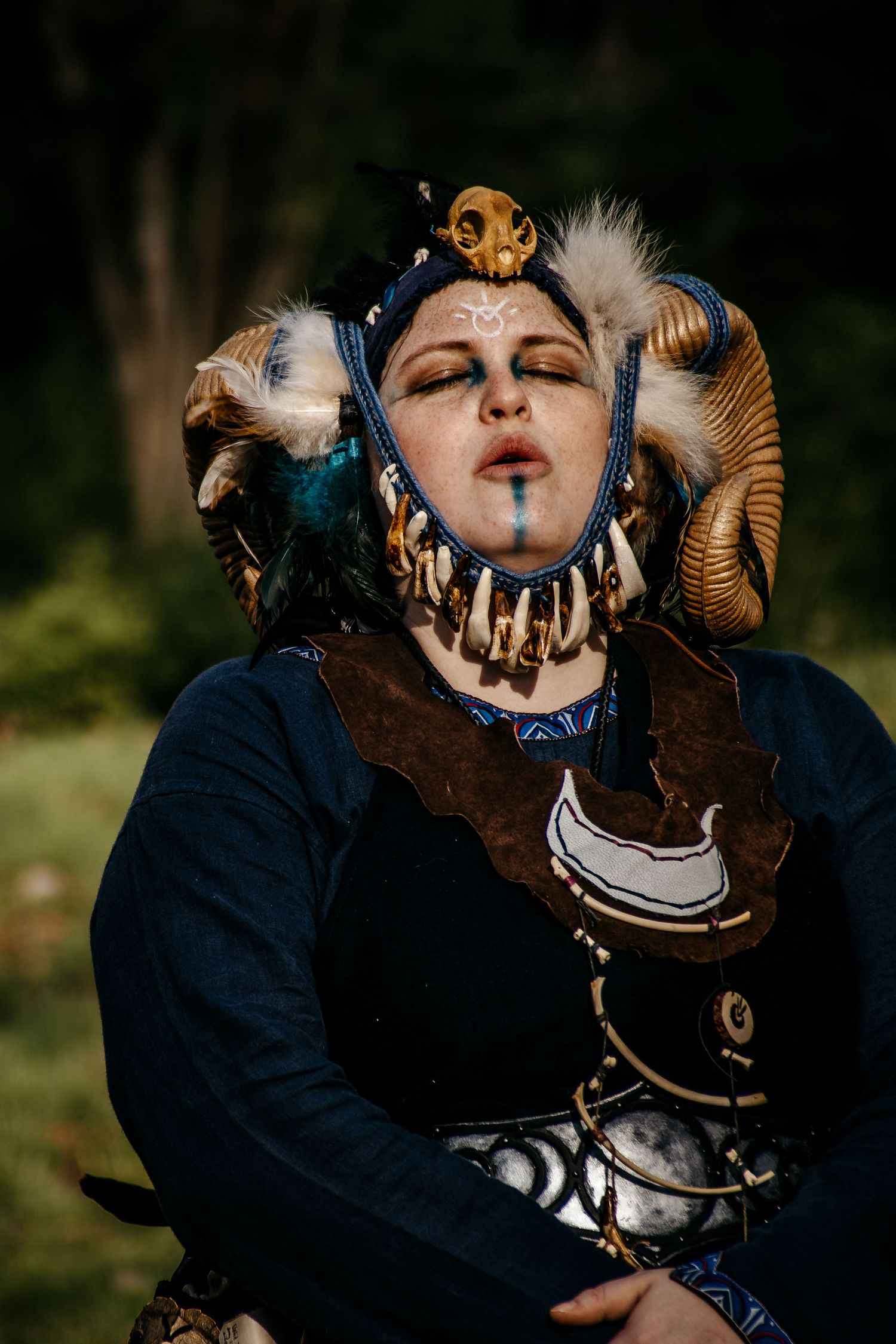 Person in elaborate fantasy costume with ram horns, feathers, bone jewelry, and face paint poses outdoors.