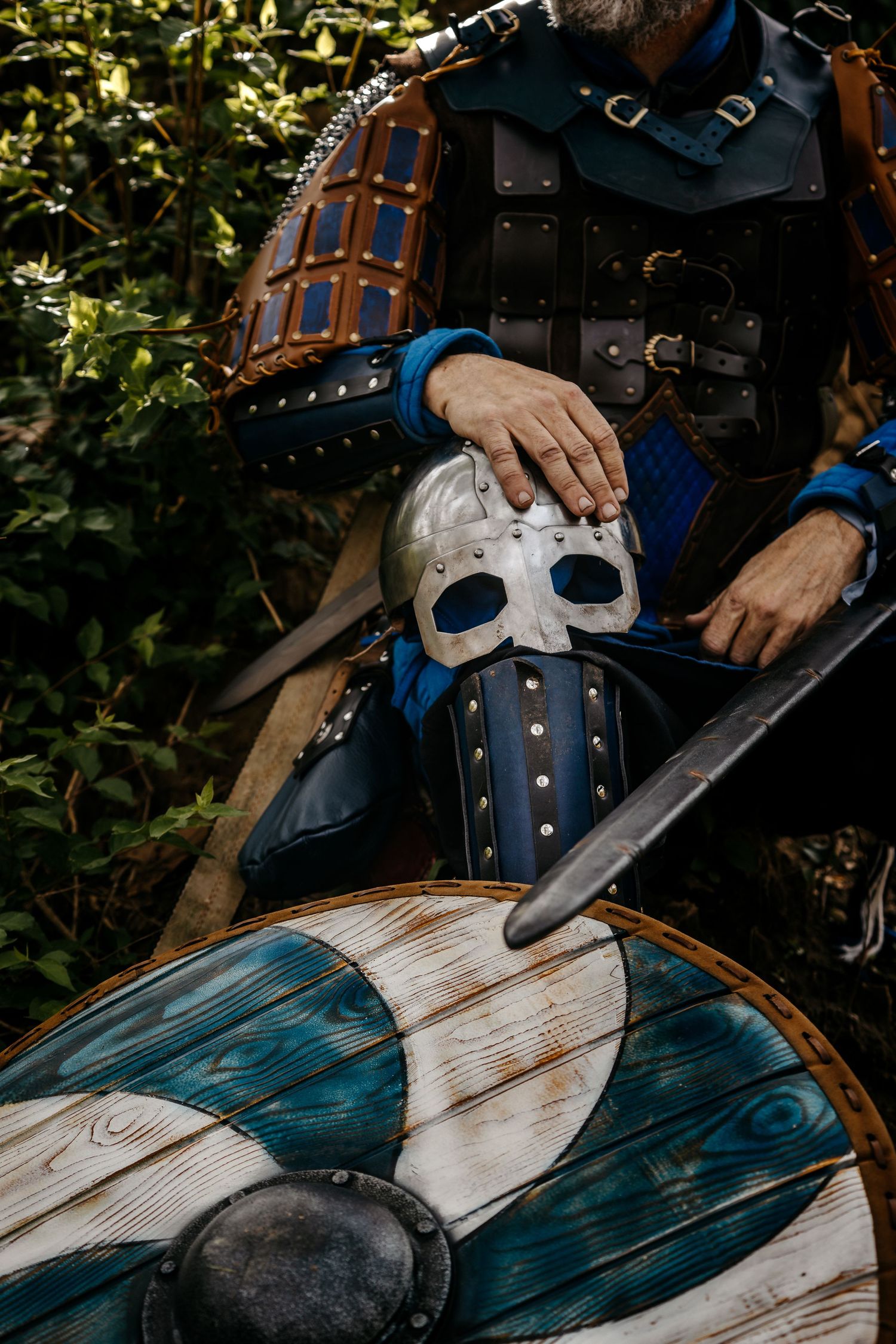 Viking warrior in leather armor holding a metal helmet and sword with a painted wooden shield.