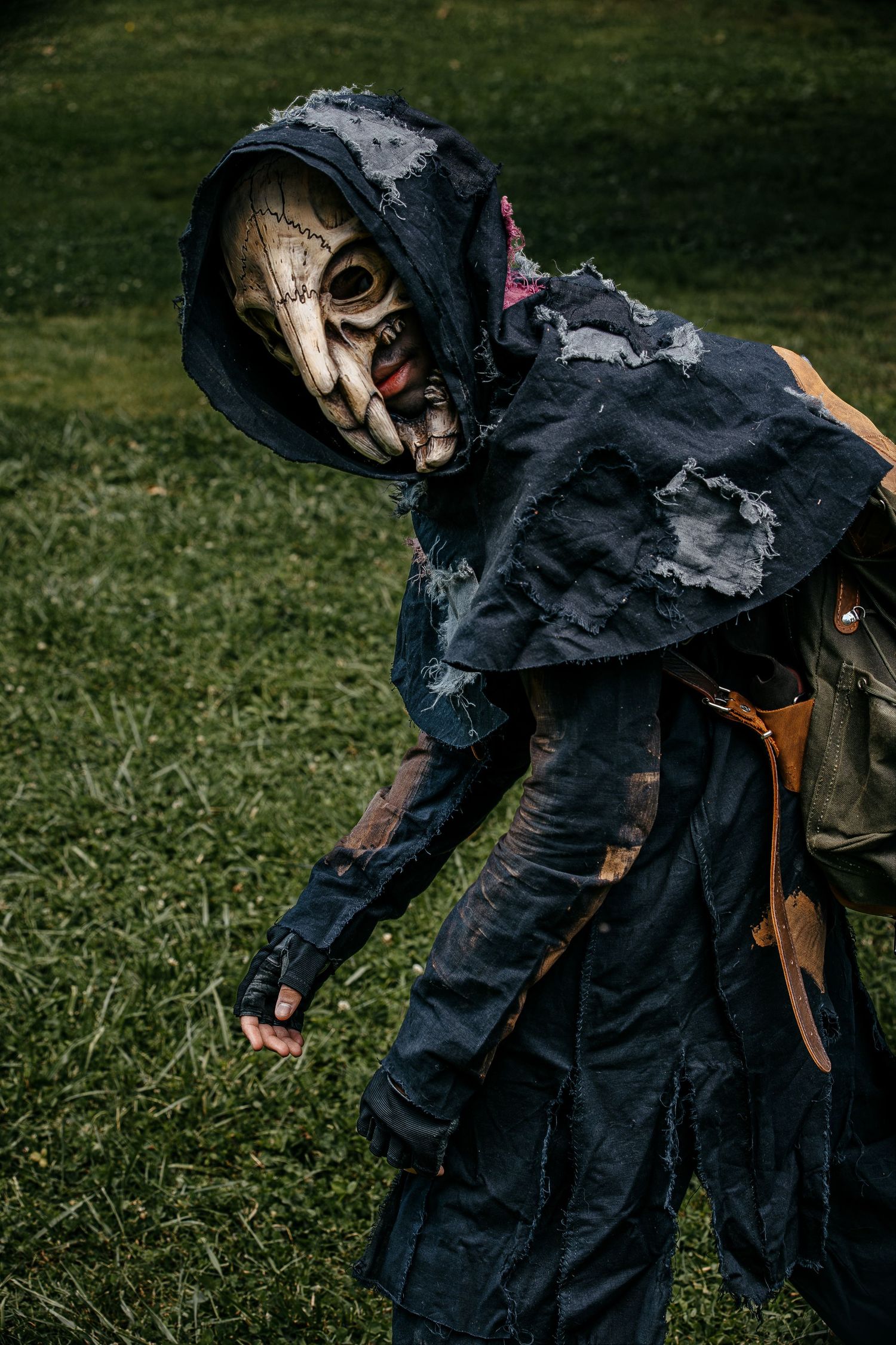 Person in tattered dark hooded cloak and skull mask costume poses outdoors on green grass for Halloween.