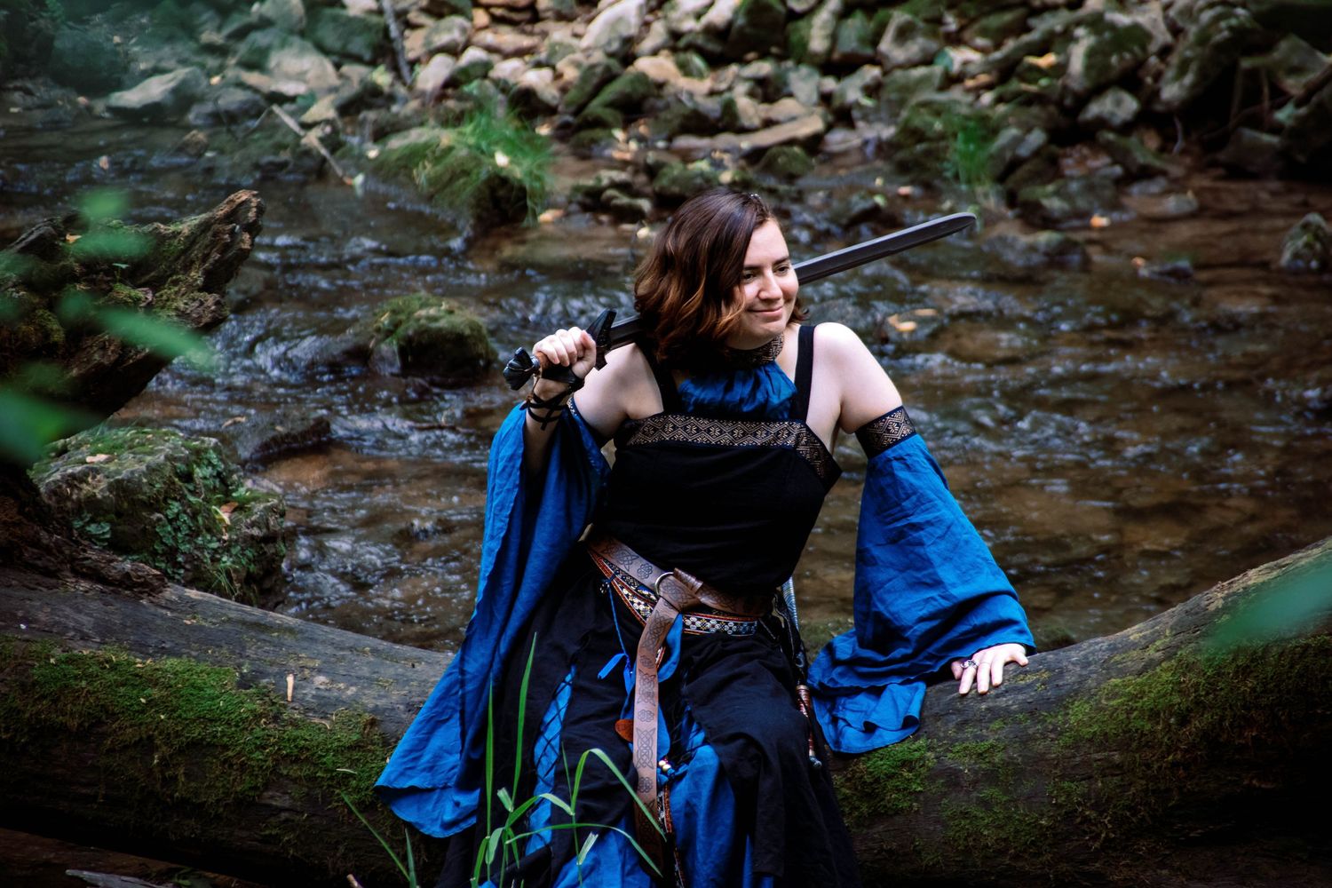 Person in blue dress poses near mossy rocks and flowing water in a forest creek setting.