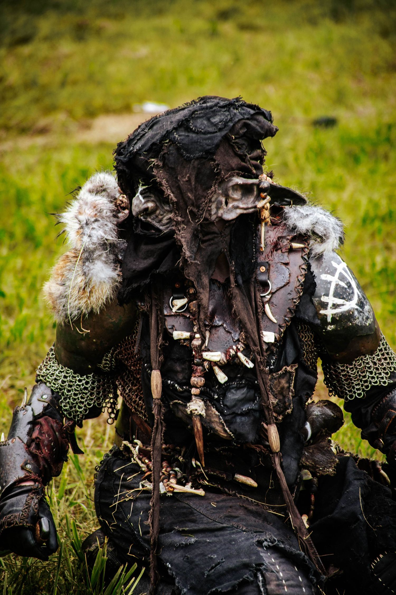 A person wearing a dark, tattered post-apocalyptic costume with fur trim and chainmail details stands in a grassy field.