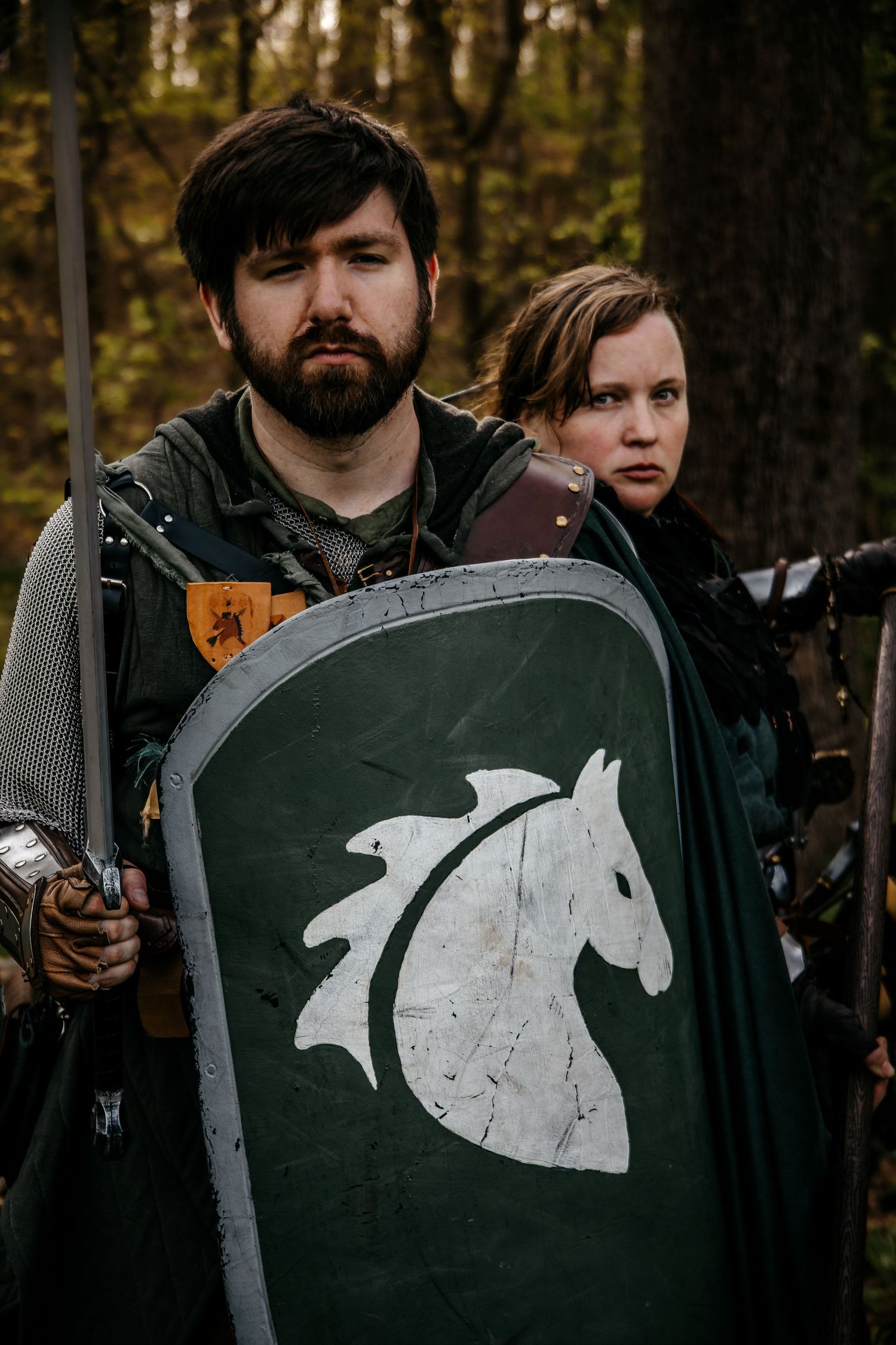 Two medieval LARP warriors in chainmail hold a green shield with white horse emblem in a forest setting.