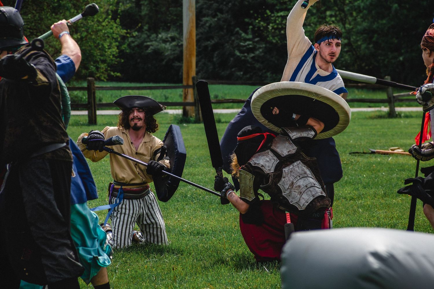 People in medieval costumes engage in a live-action roleplay battle with swords and shields on a grassy field.