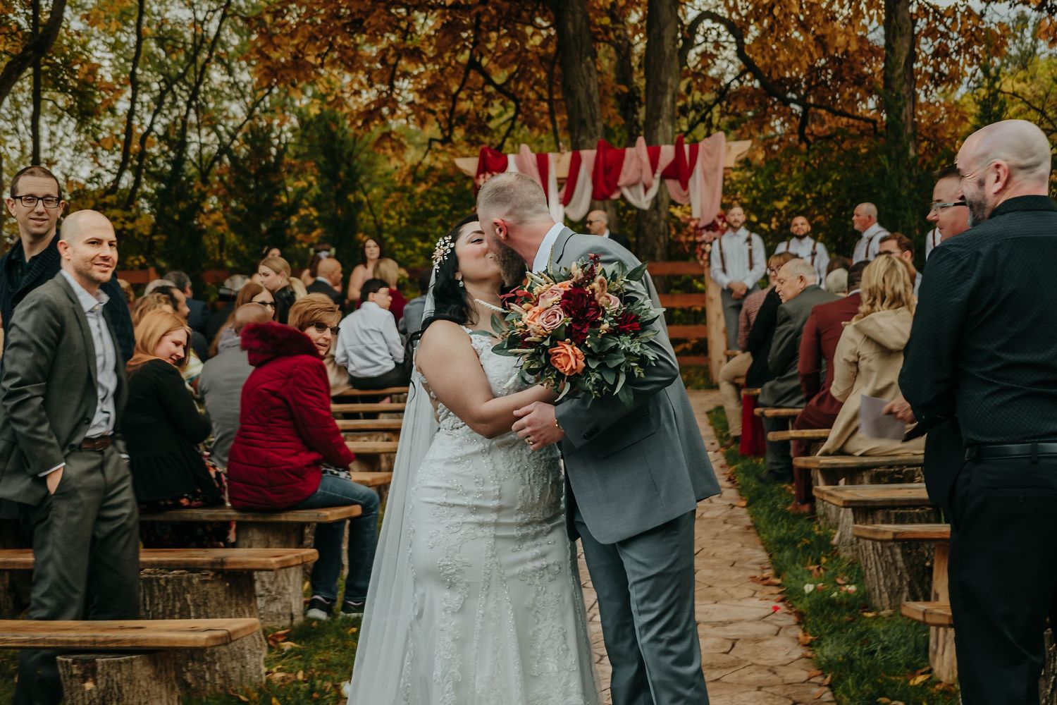 A couple shares their first kiss during an autumn outdoor wedding ceremony with wooden benches and fall foliage.