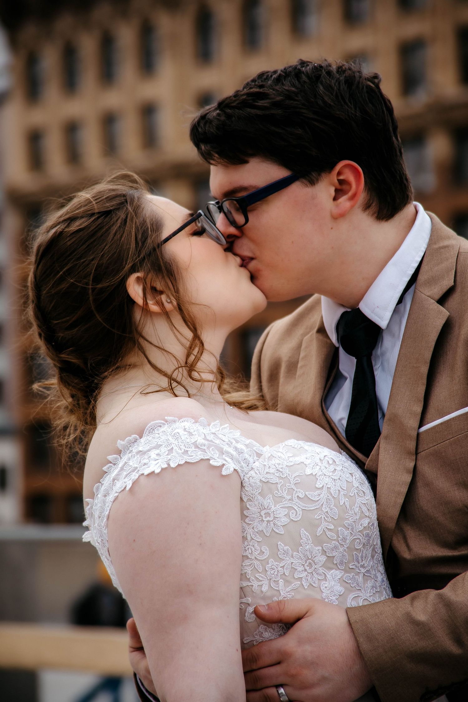 A romantic wedding couple share a tender kiss, with the bride in lace and groom in tan suit against an urban backdrop.