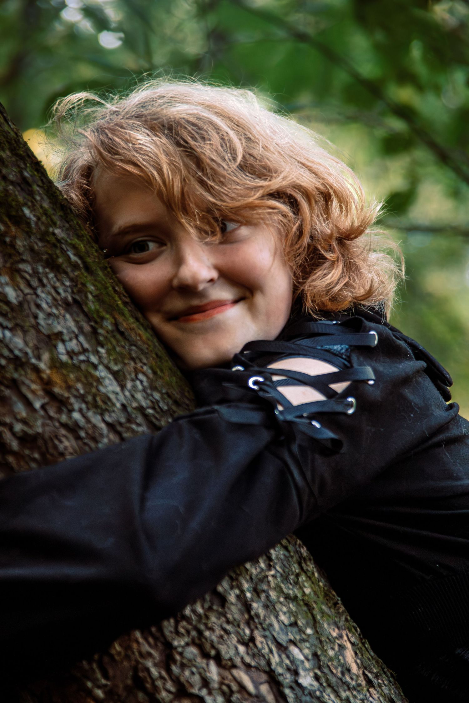 A person with curly blonde hair wearing a black lace-up sweater leans against a tree trunk in a natural outdoor setting.
