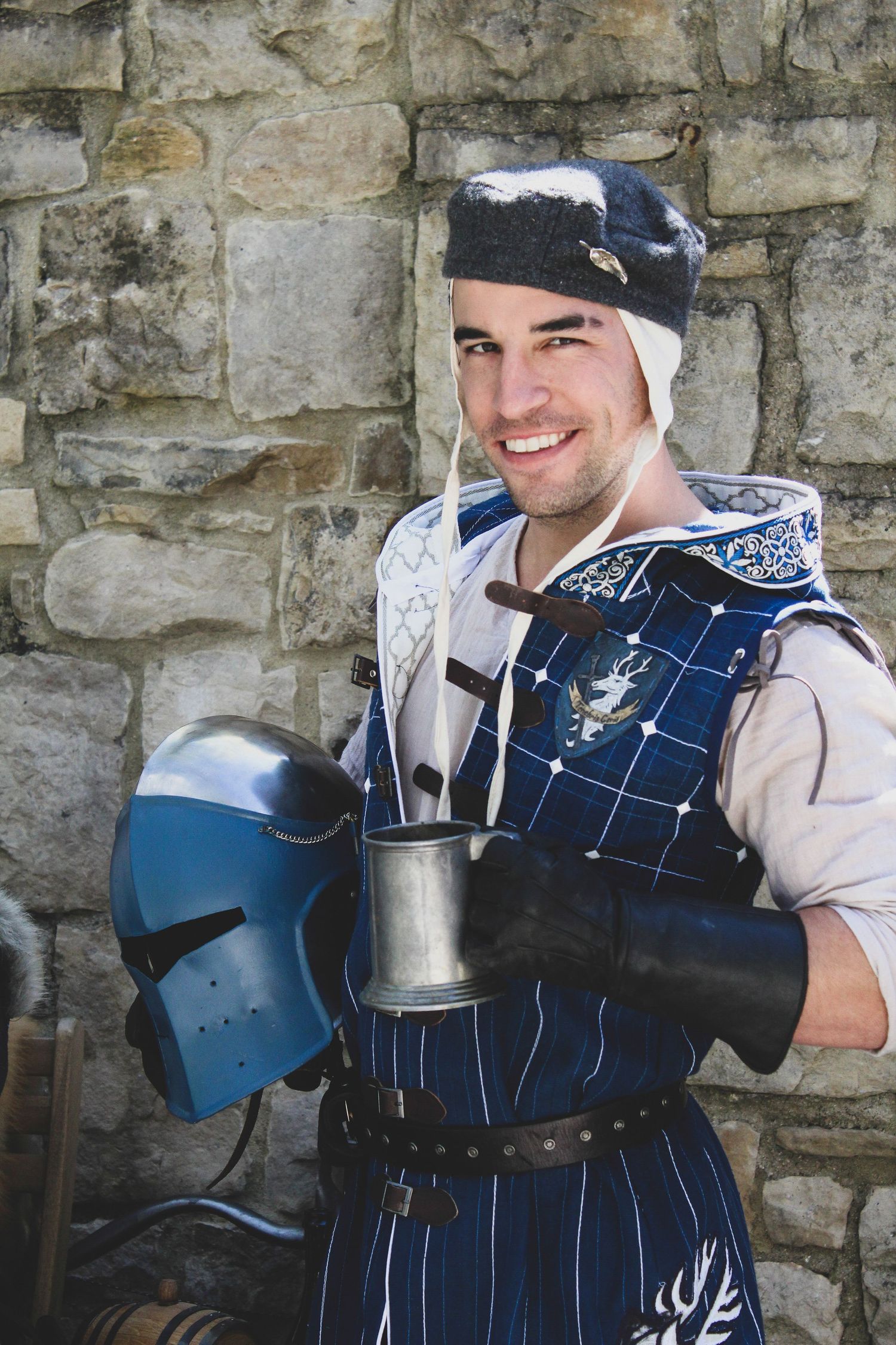 Medieval knight wearing blue and white striped armor with metal shoulder plates poses against stone wall.