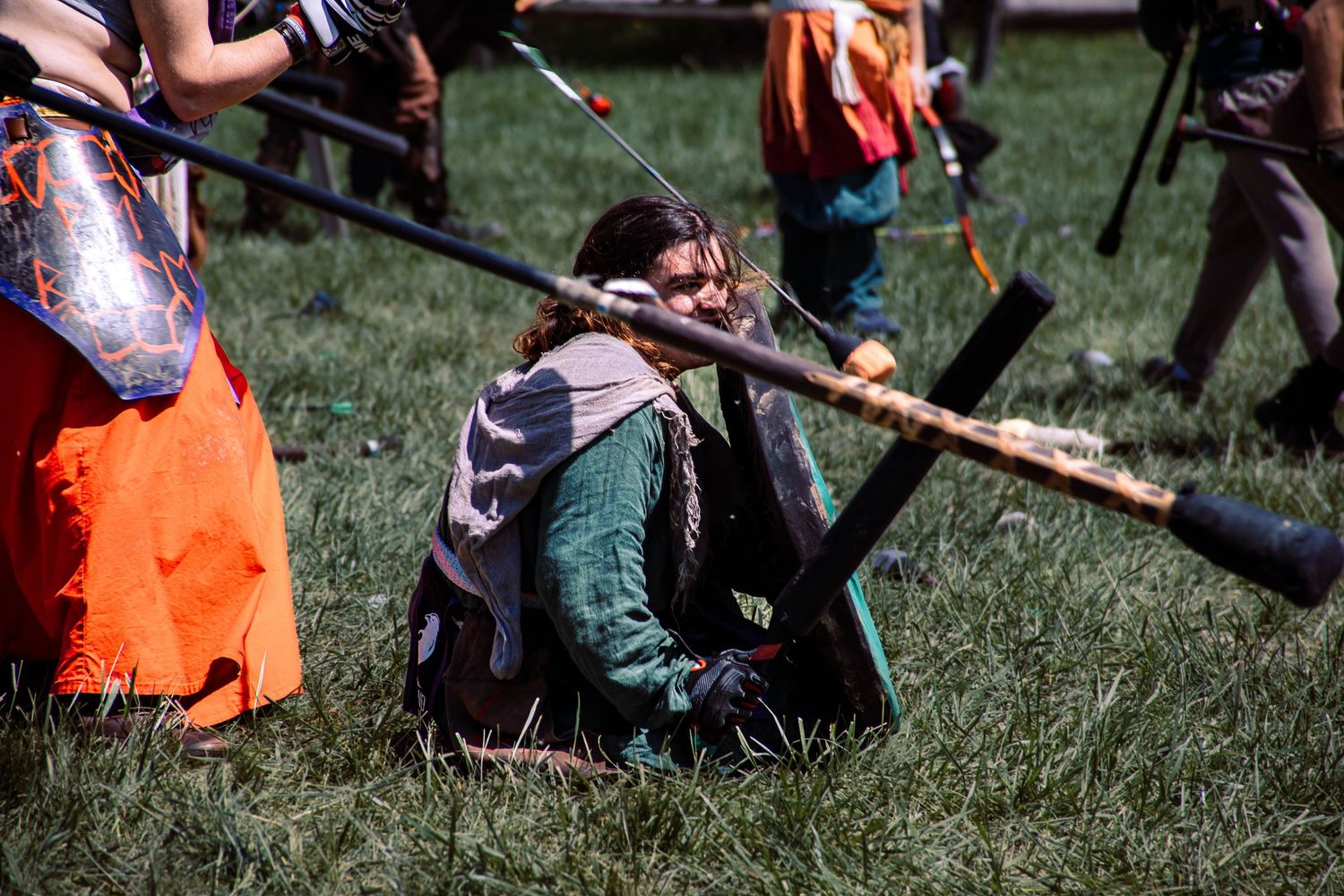 Medieval reenactor in green tunic kneels in grass while holding wooden staff during historical demonstration.