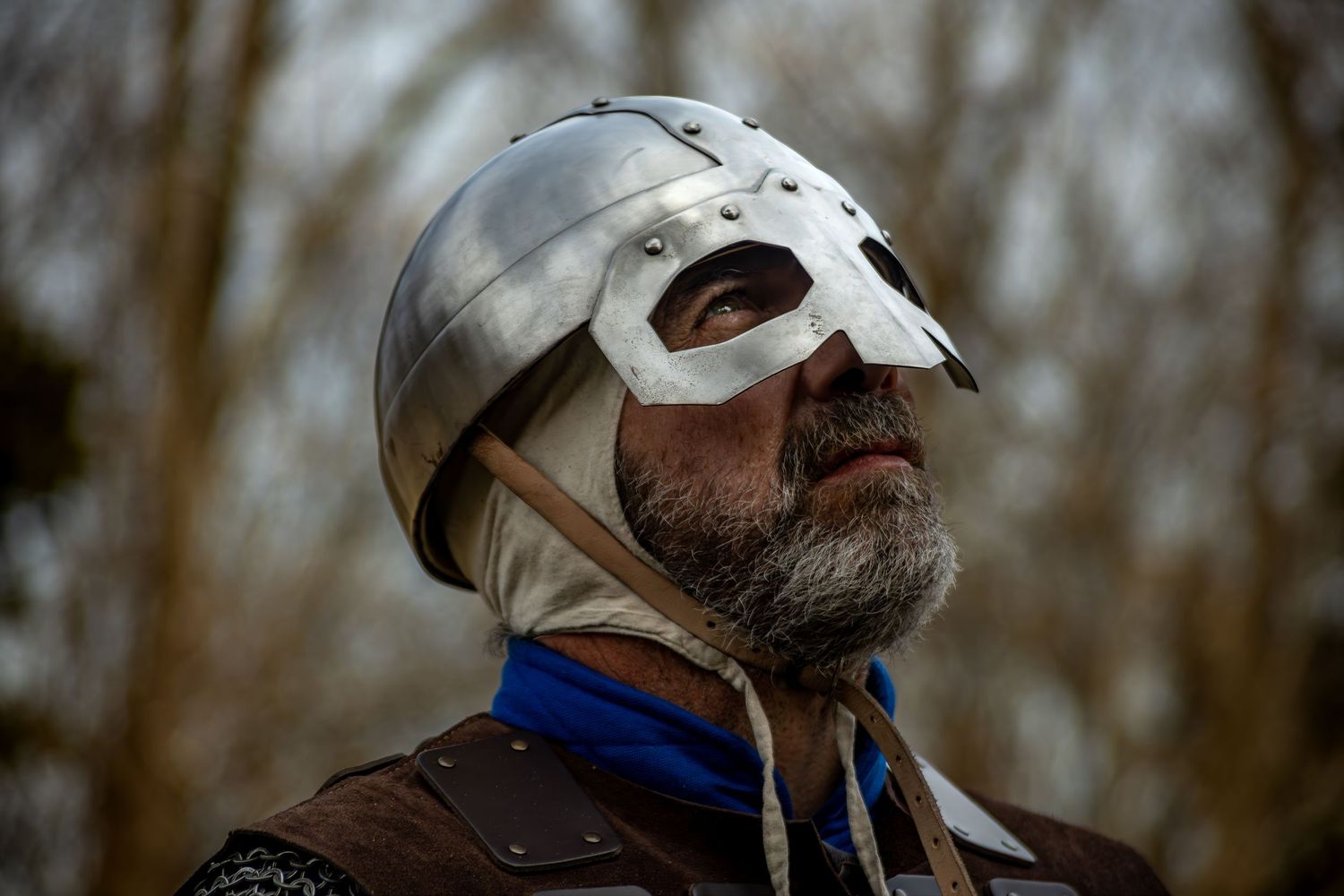 Bearded man wearing a medieval knight helmet and armor, looking upward against a blurred winter trees background.