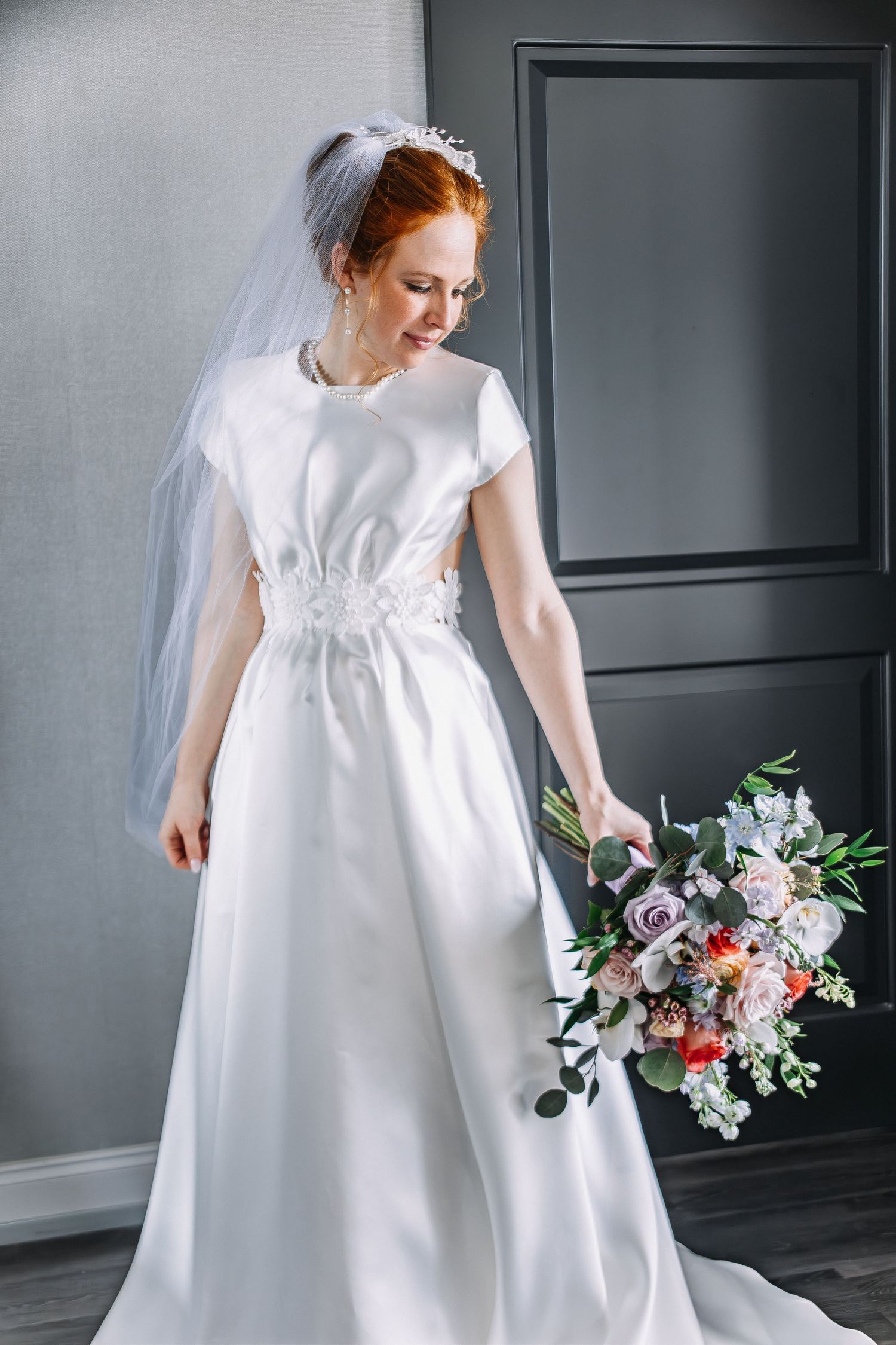 Red-haired bride in white satin gown with veil holds colorful bouquet, smiling softly in elegant gray room.