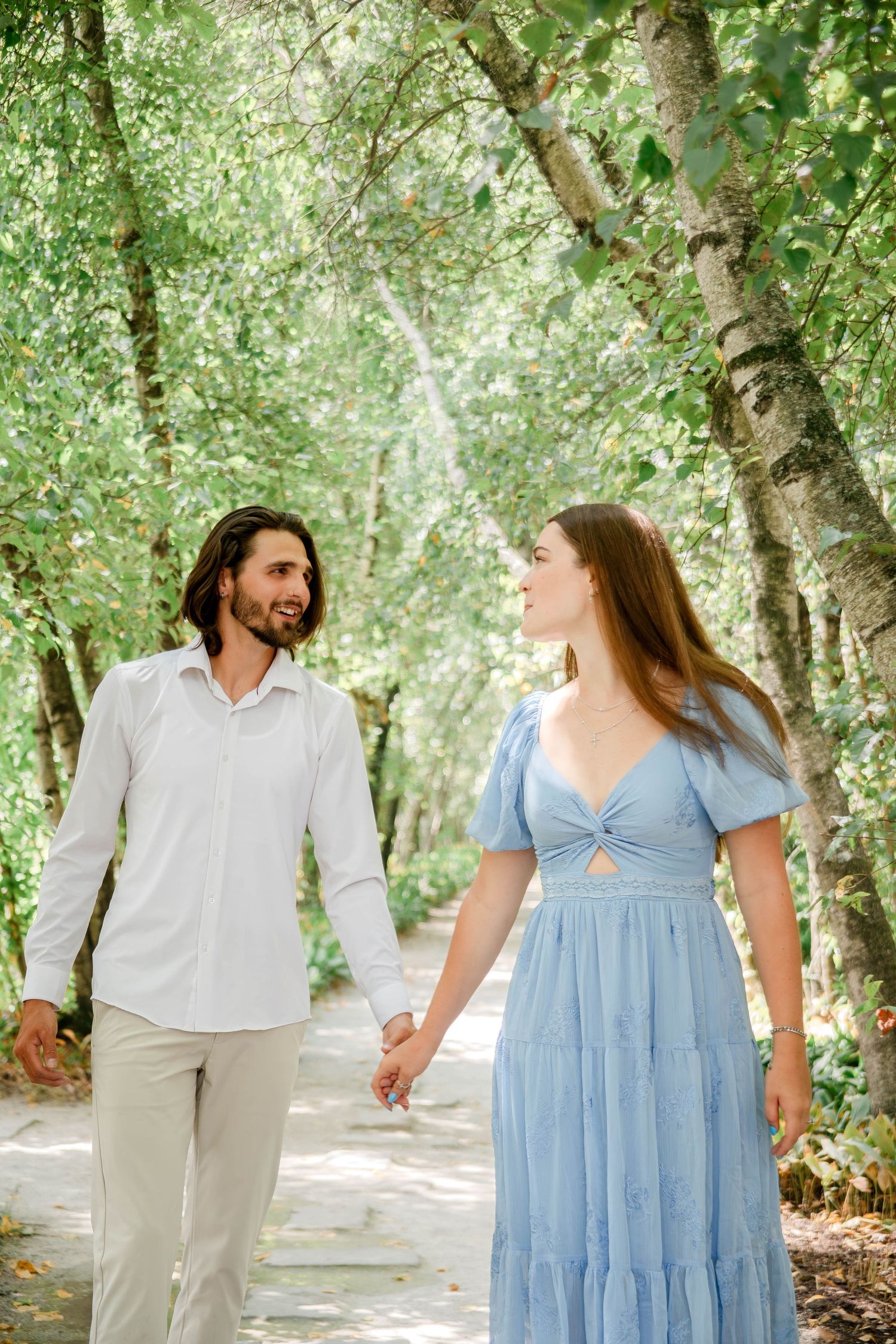 A couple strolls hand in hand along a tree-lined path in summer attire.