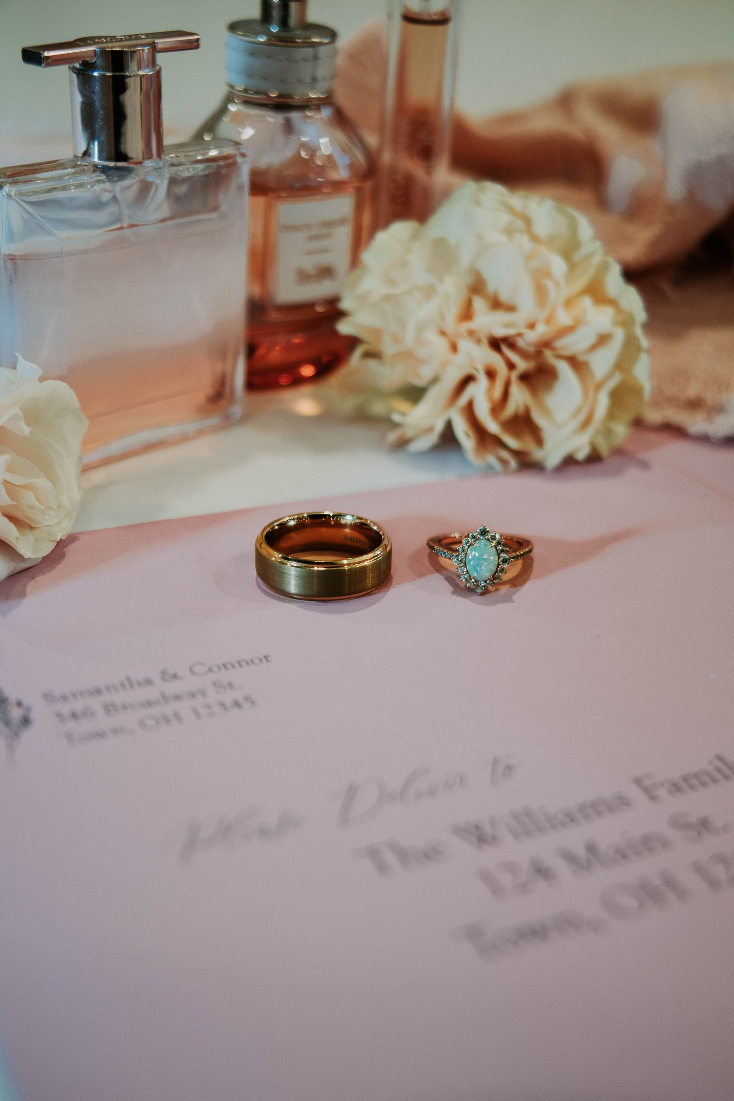 Gold wedding band and oval turquoise engagement ring rest on pink envelope beside perfume bottles and white flowers.