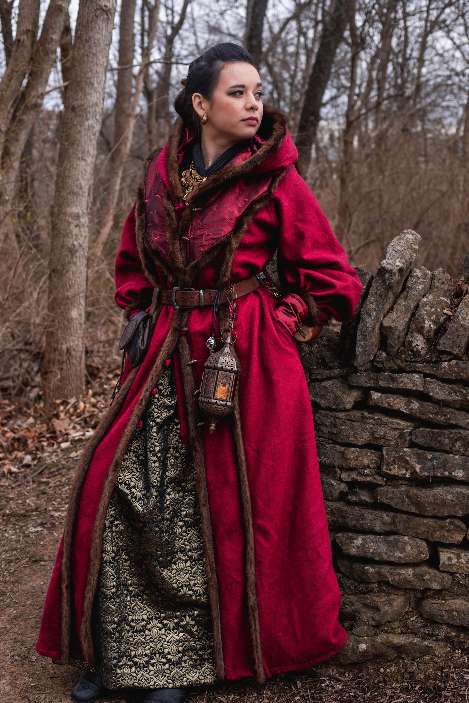 A dramatic red hooded cloak worn in a forest setting against a stone wall backdrop.