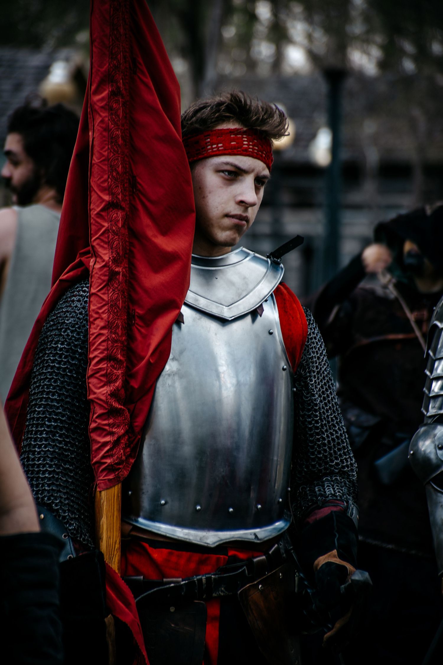 Young man in medieval knight armor holding a red banner at a historical reenactment event.