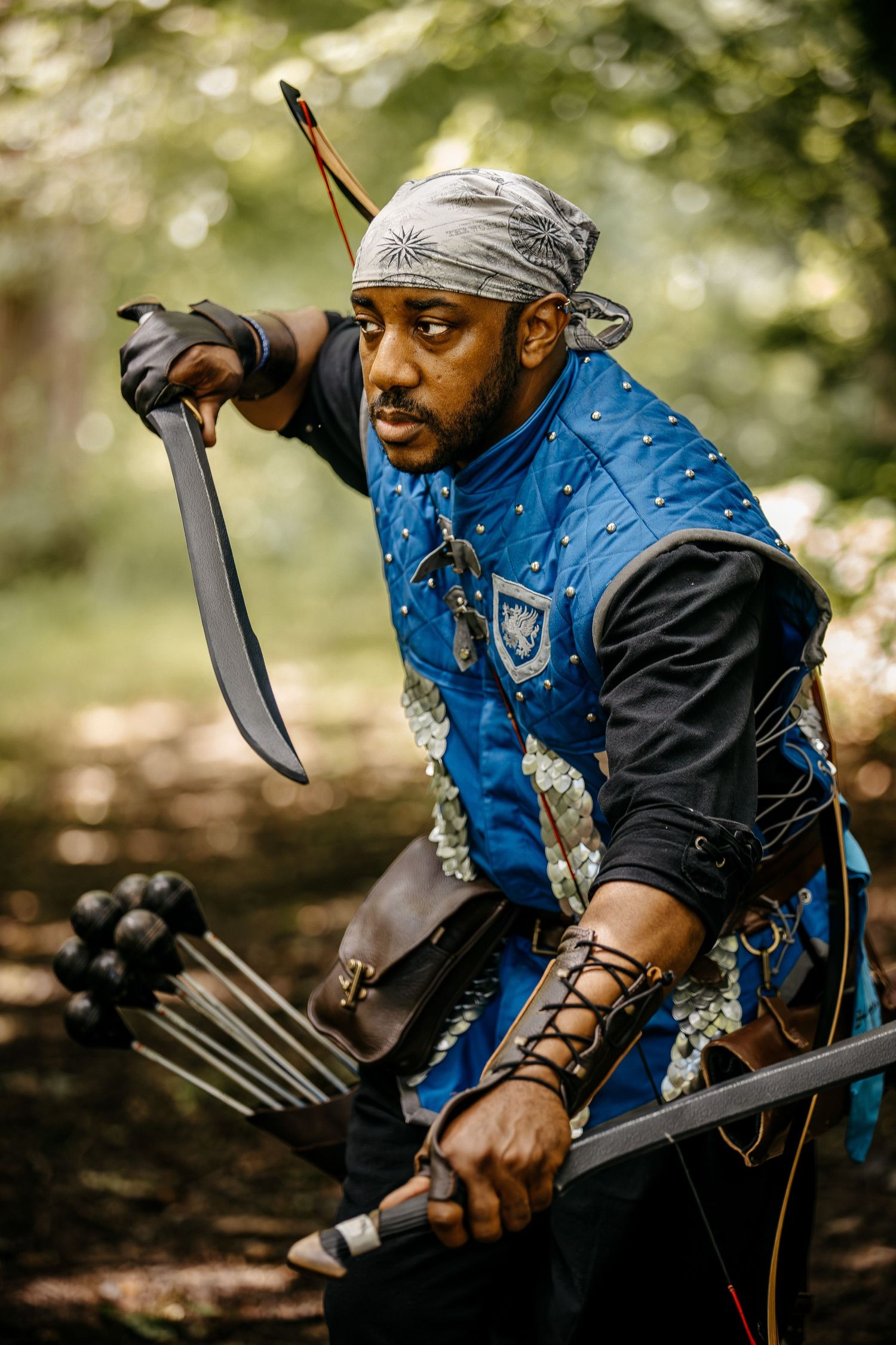 A Black man in medieval archer costume with blue vest, bandana, holding a sword and bow in a forest setting.