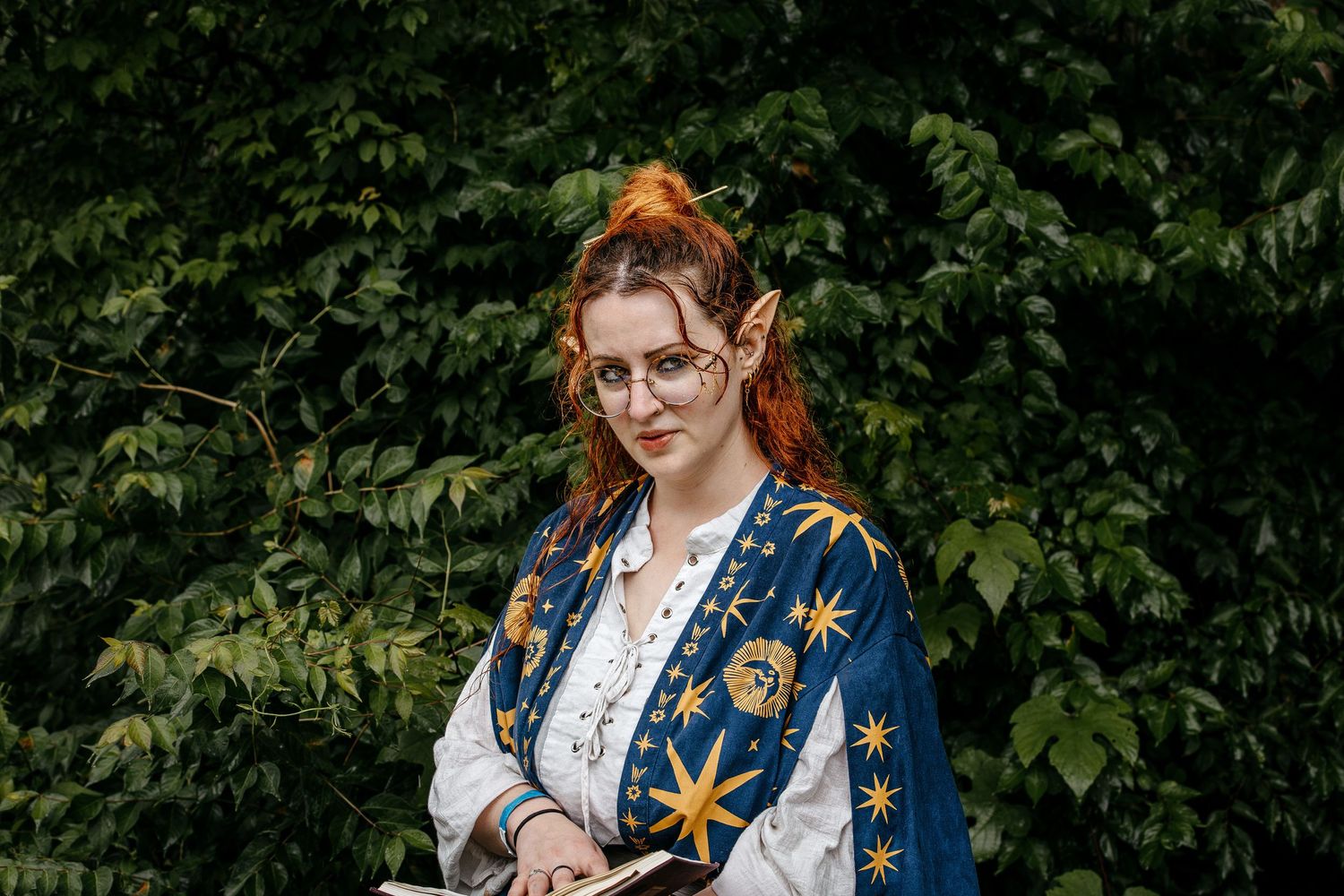 Woman in celestial elf costume with pointed ears, red hair, and star-patterned robe holding a book outdoors.