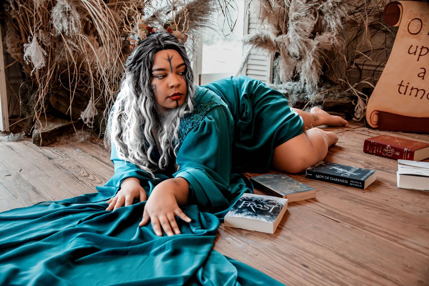 Woman in teal robe with silver braided hair poses on wooden floor surrounded by books in fantasy-themed setting.