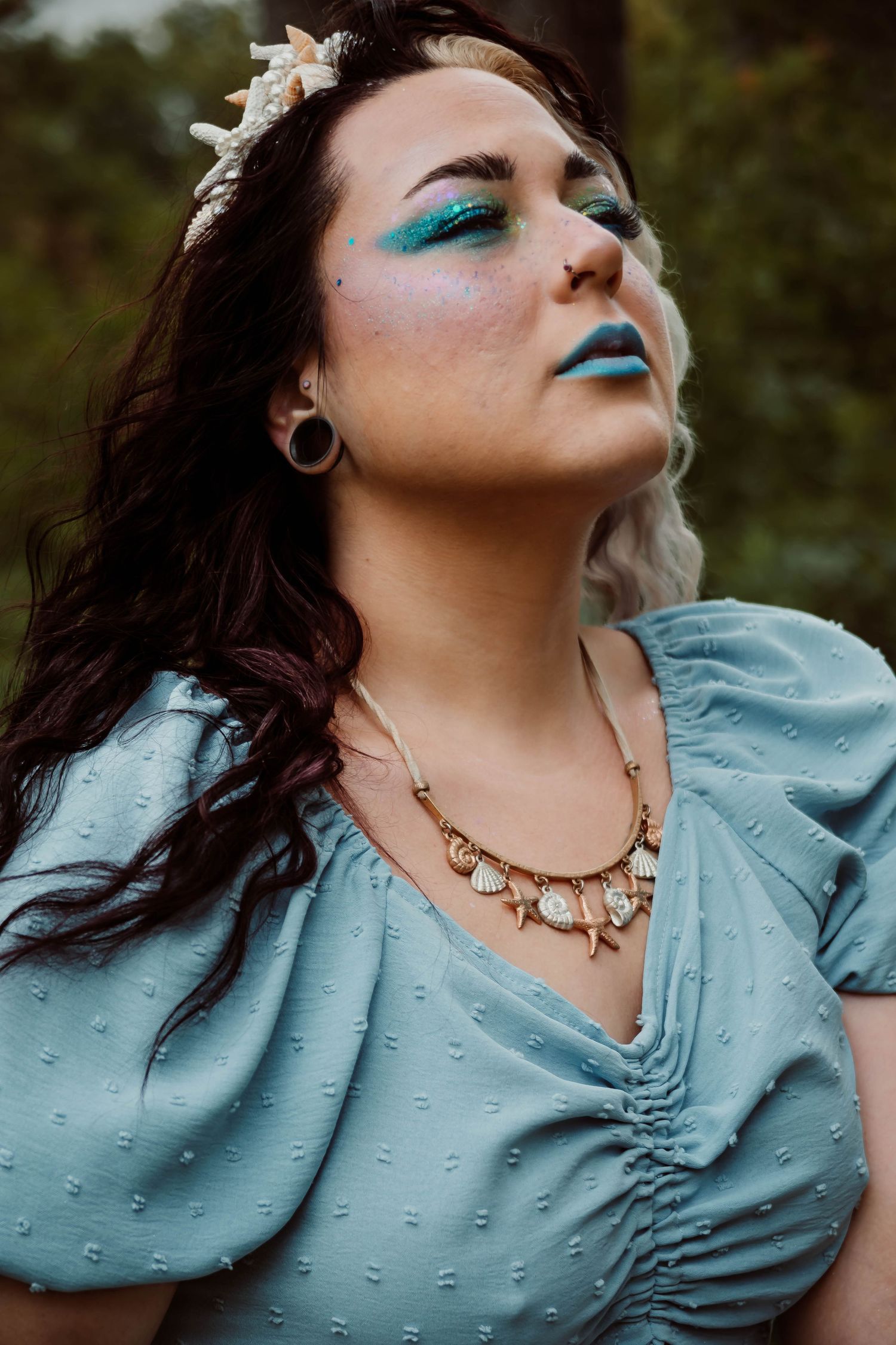 Woman in mermaid costume with teal eye makeup, shell crown, and seashell necklace wearing a light blue dress outdoors.