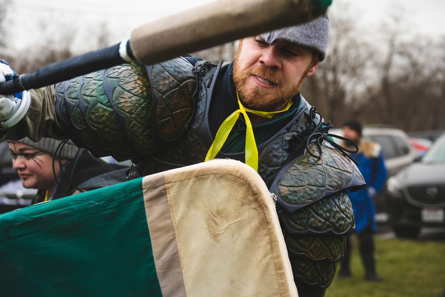 A person wearing medieval armor and a yellow scarf stands ready with a weapon during an outdoor historical reenactment.