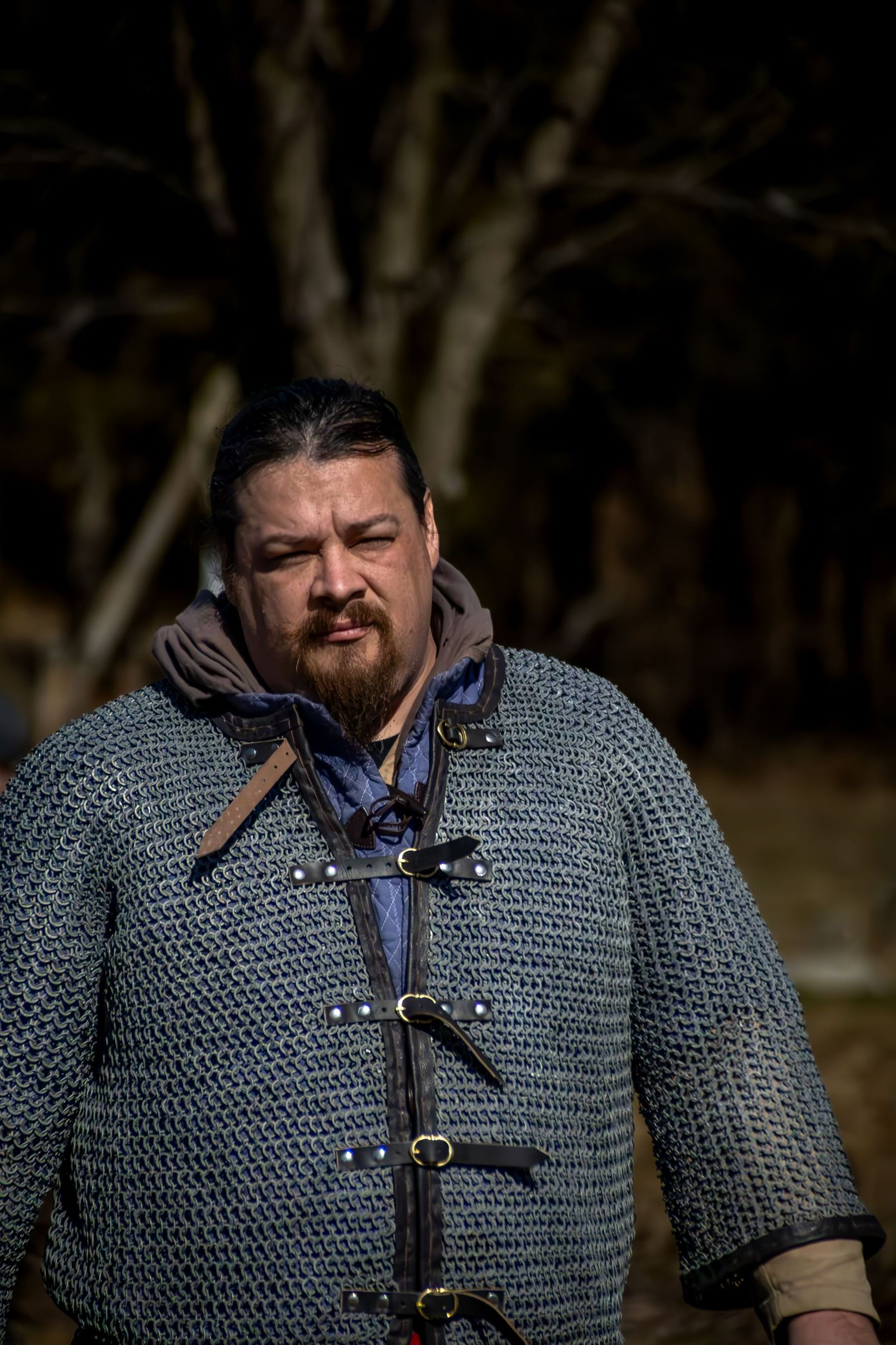 Medieval reenactor wearing chainmail armor and leather buckles stands in front of dark trees.