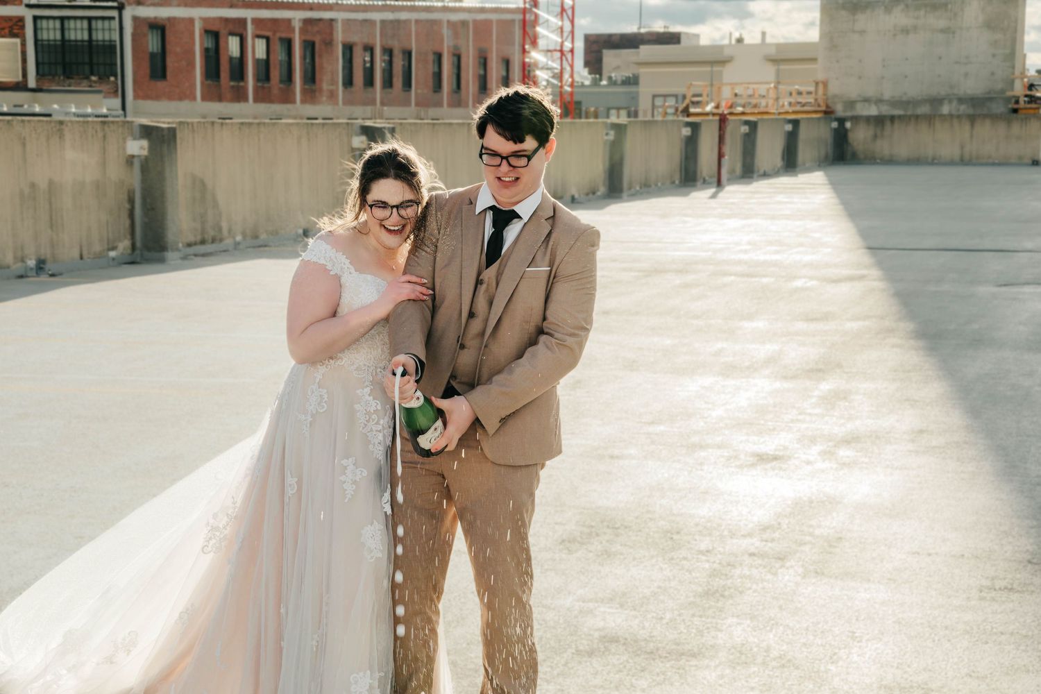 A couple in wedding attire pose for photos in an urban setting with brick buildings and industrial architecture.