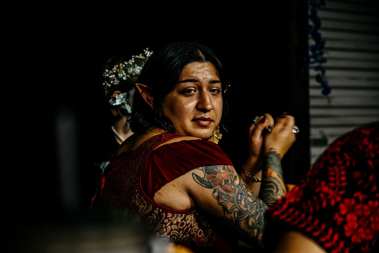 A tattooed South Asian woman in a red saree with floral hair accessories looks emotional during a wedding ceremony.