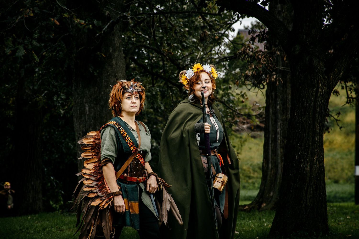 Two women in fantasy medieval costumes pose outdoors among trees in a lush green park setting.