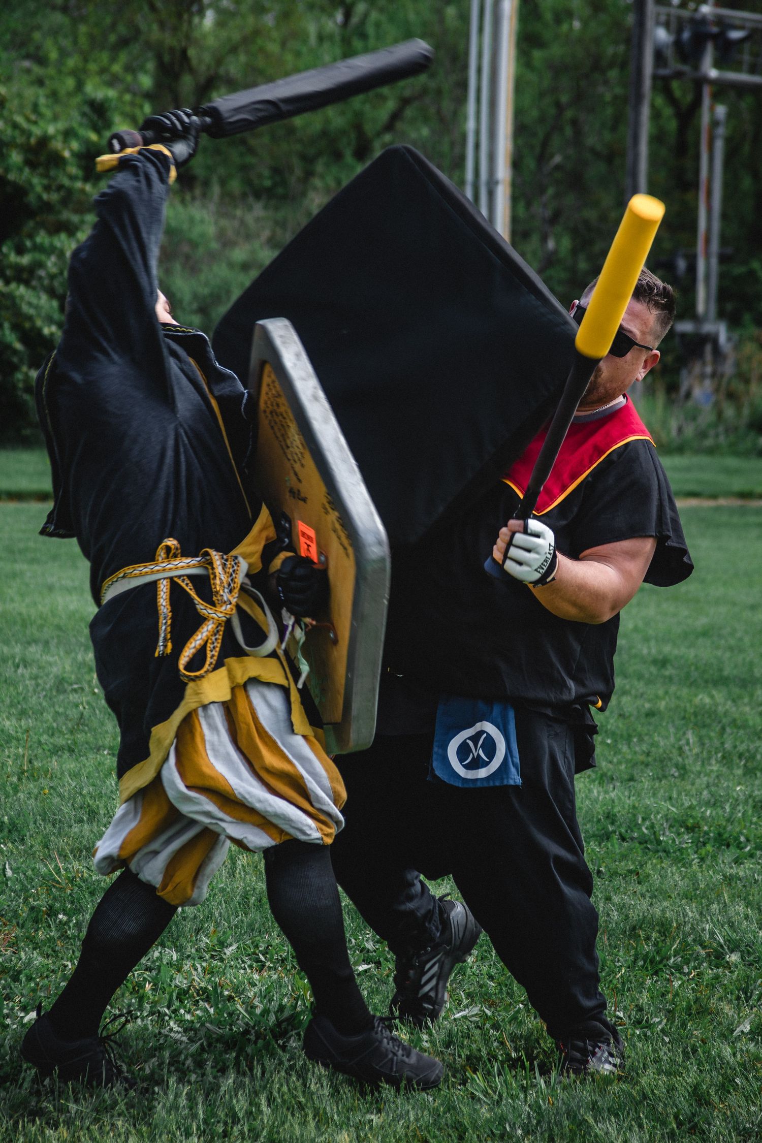Two costumed fighters battle with foam swords and shields on grass at an outdoor medieval combat event.