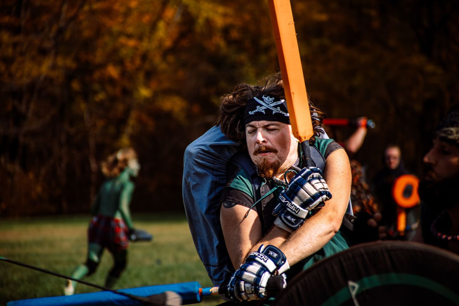 A bearded man in medieval gear wields an orange weapon during an outdoor LARP battle event in autumn woods.