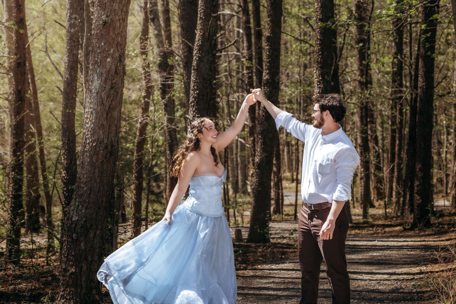 Couple dancing in sunlit forest, bride in blue gown twirling with groom in white shirt and brown pants.