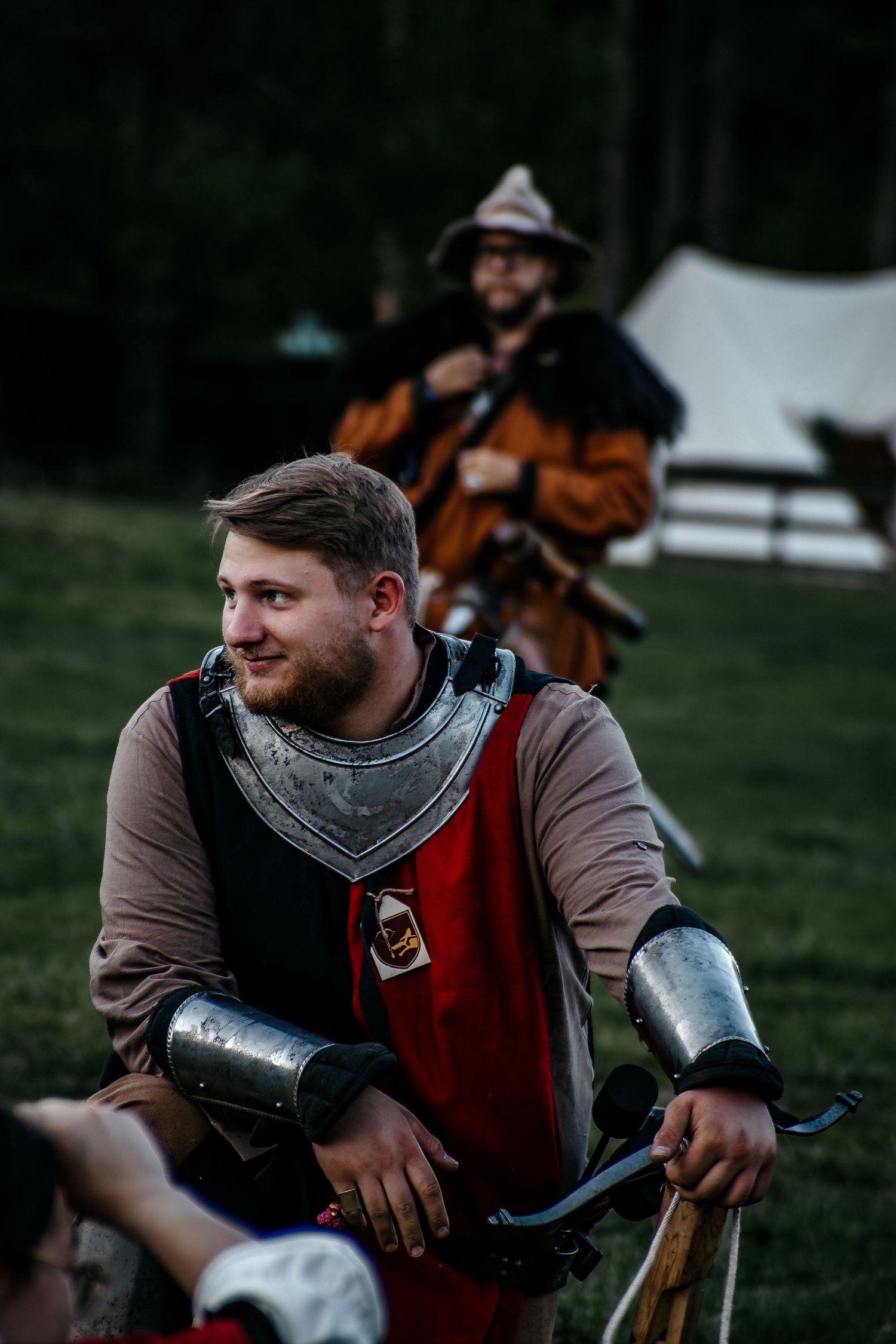 Man in medieval knight armor at historical reenactment event, with another costumed participant visible in background.