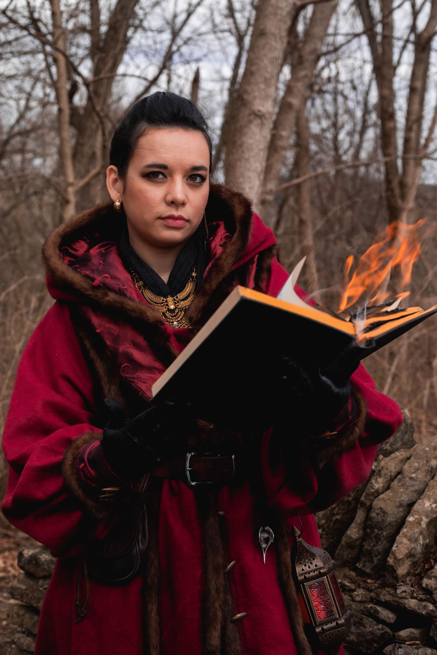 A person in a burgundy winter cloak stands in a forest setting with flames visible in the background.