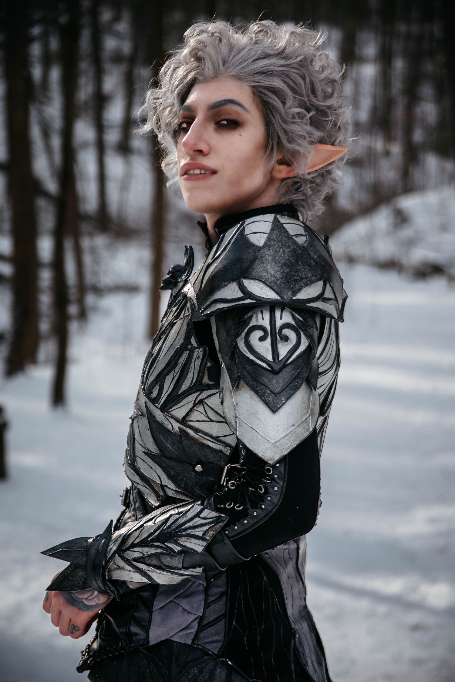 A cosplay model in a black and silver armored costume poses dramatically in a snowy winter forest setting.