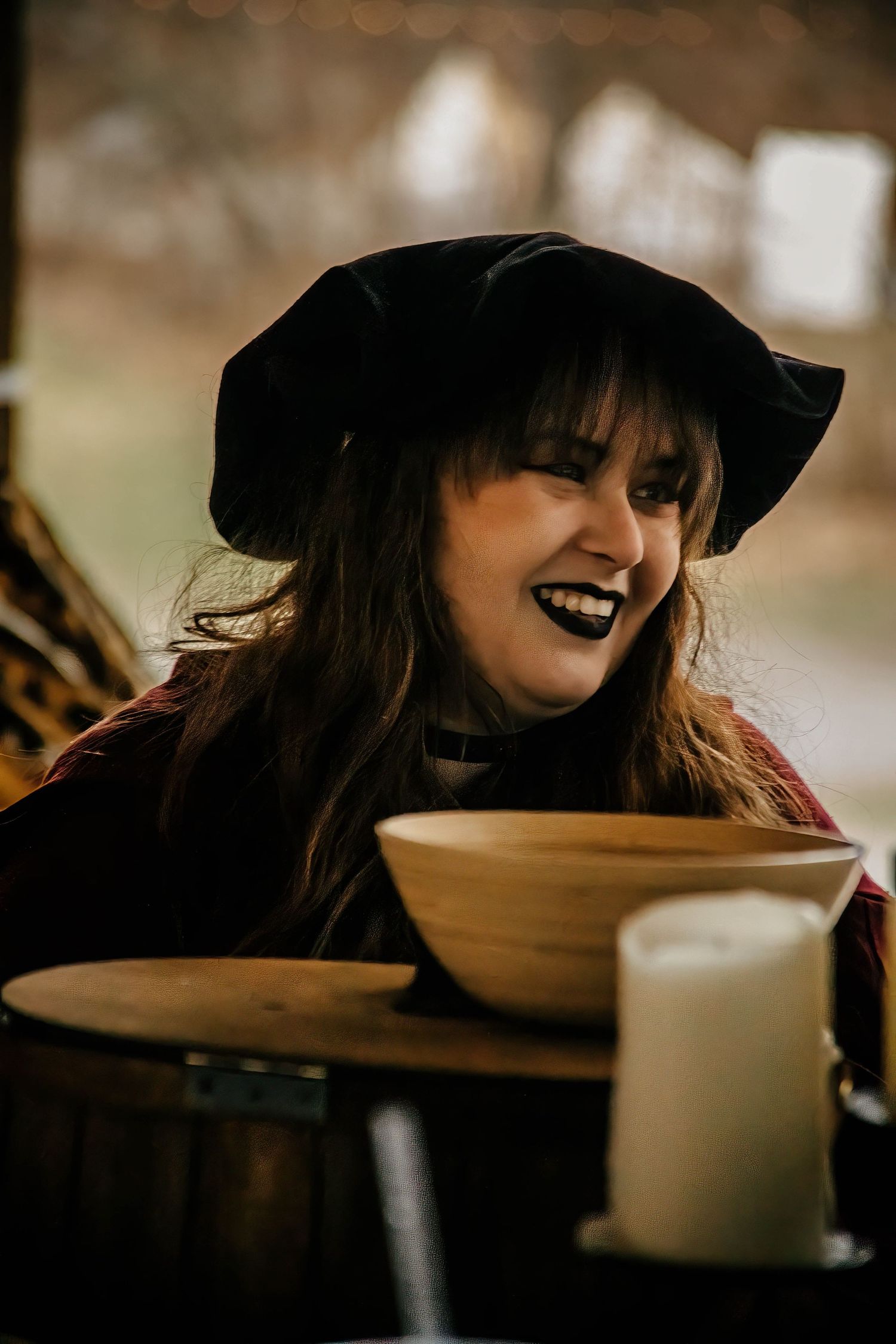 A figure in a black witch hat leans over a table with candles in moody lighting.