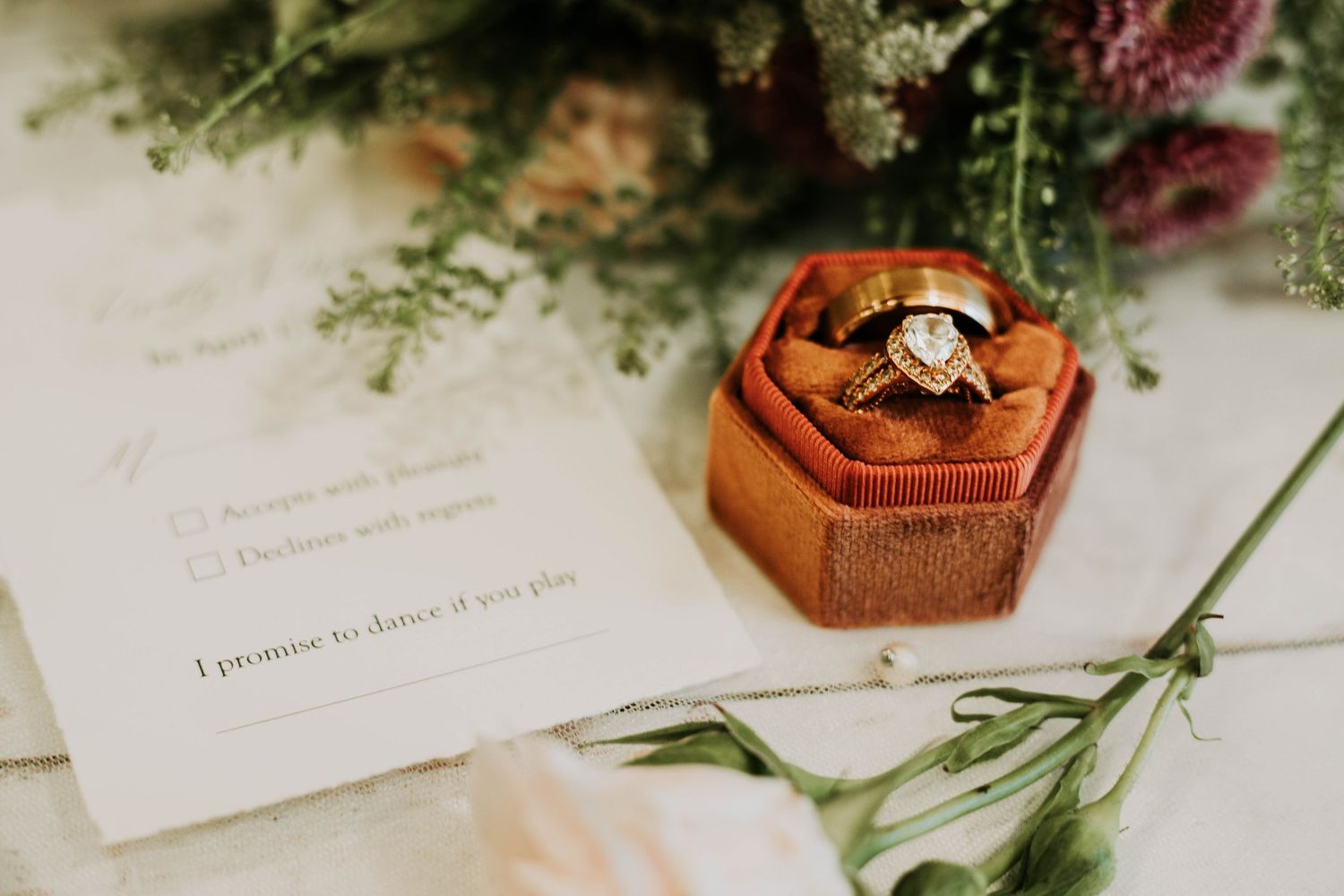 Wedding rings displayed in a wooden hexagonal box with greenery and invitation details.