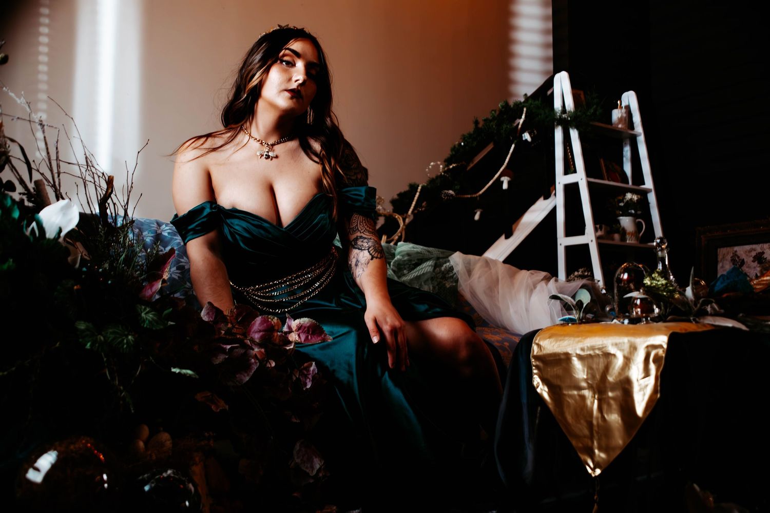 A woman in an elegant dark green off-shoulder gown poses dramatically among lush floral arrangements in moody studio lighting.