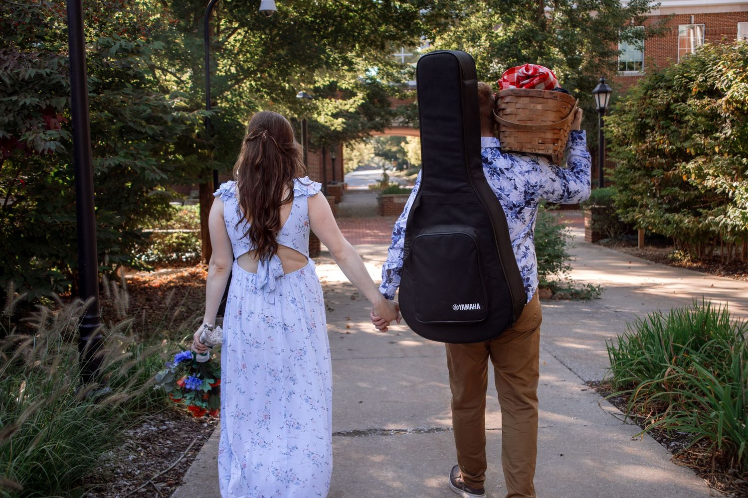 A couple walks together through a lush garden pathway surrounded by green foliage and brick walls.